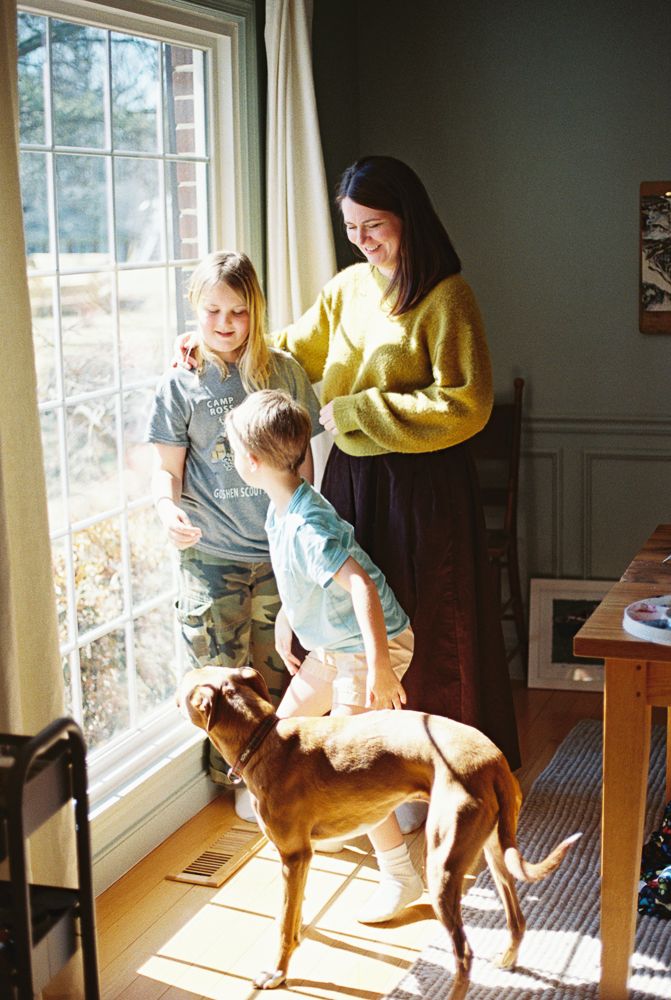 Woman in yellow sweater smiling with two children and a brown dog by a sunny window indoors.