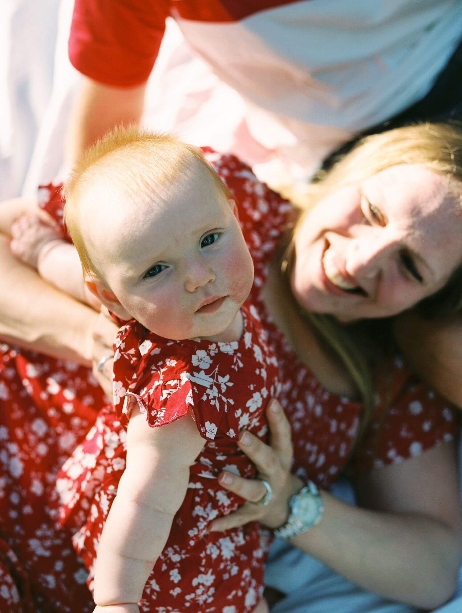 A baby in a red and white polka dot dress lies on white bedding while playing.