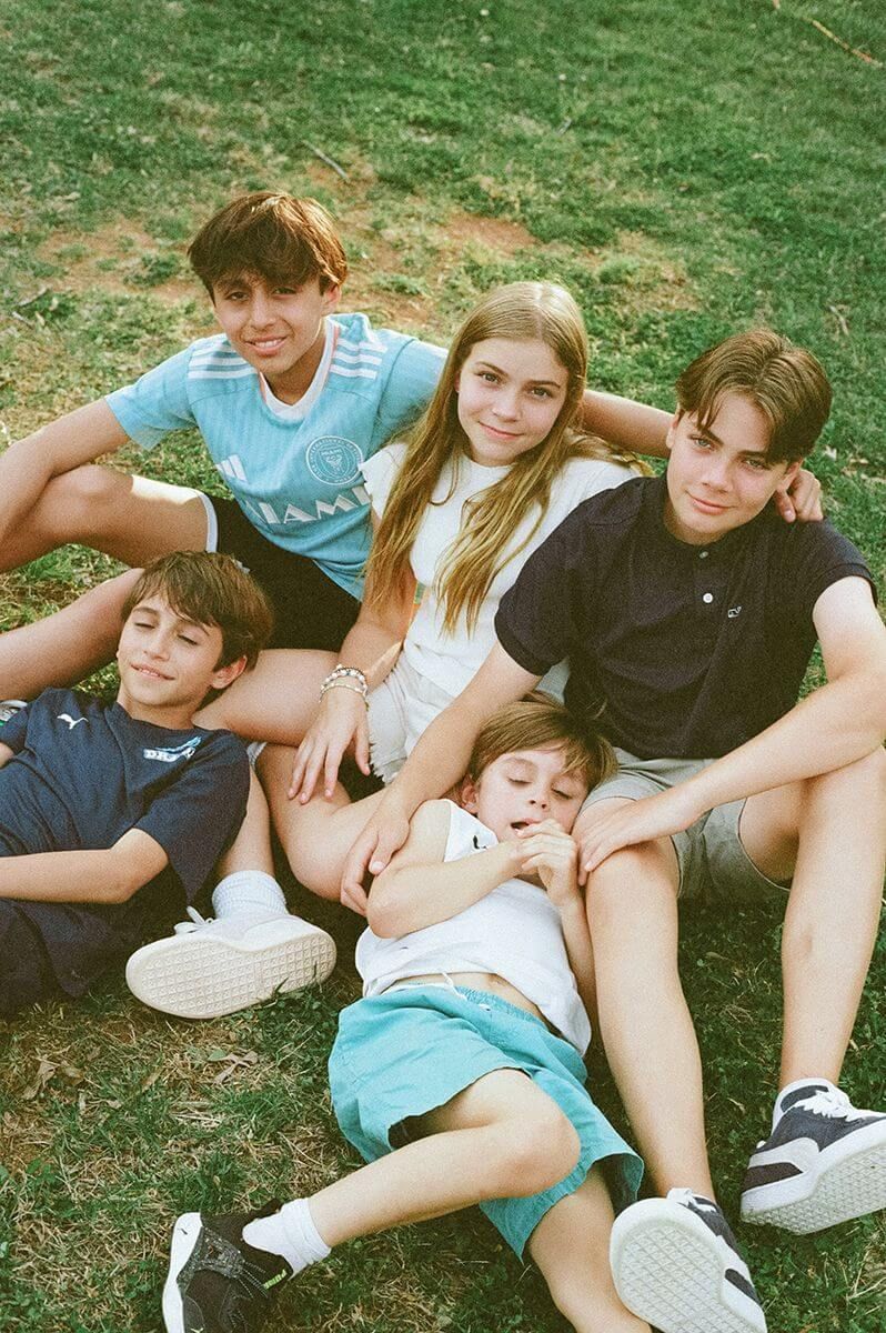 Five young people relaxing together on grass during a sunny summer day, wearing casual clothes and sneakers.