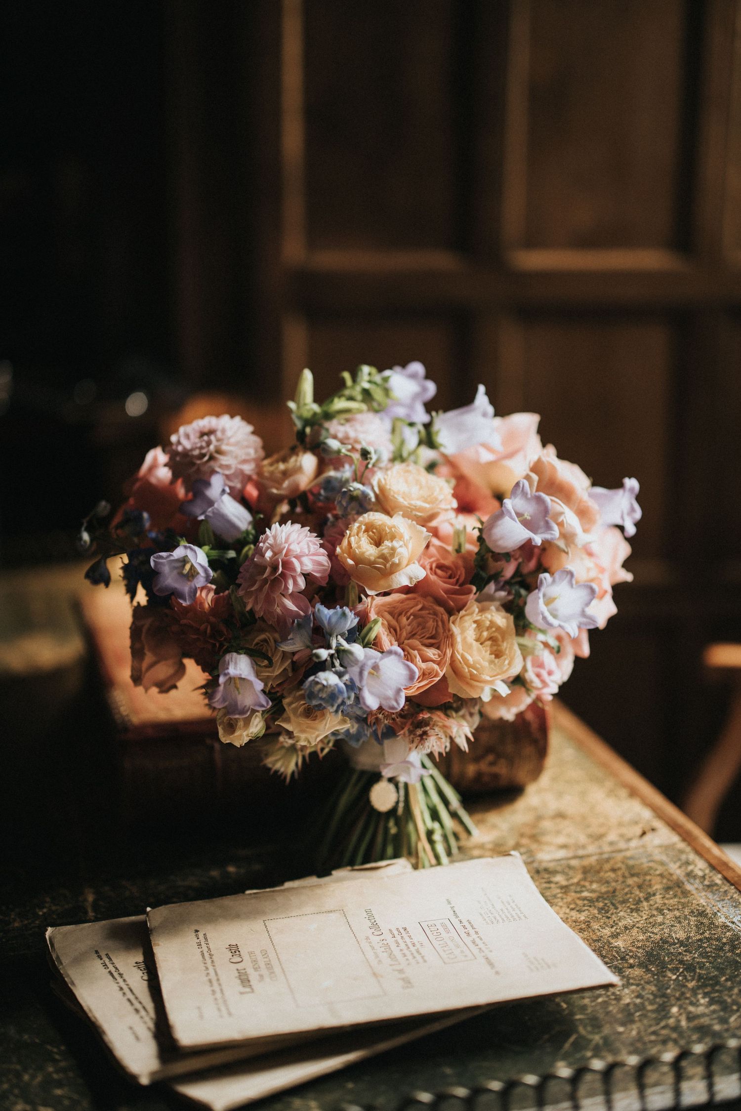 A romantic floral bouquet with pink and peach roses rests beside vintage paper on an antique table in moody lighting.
