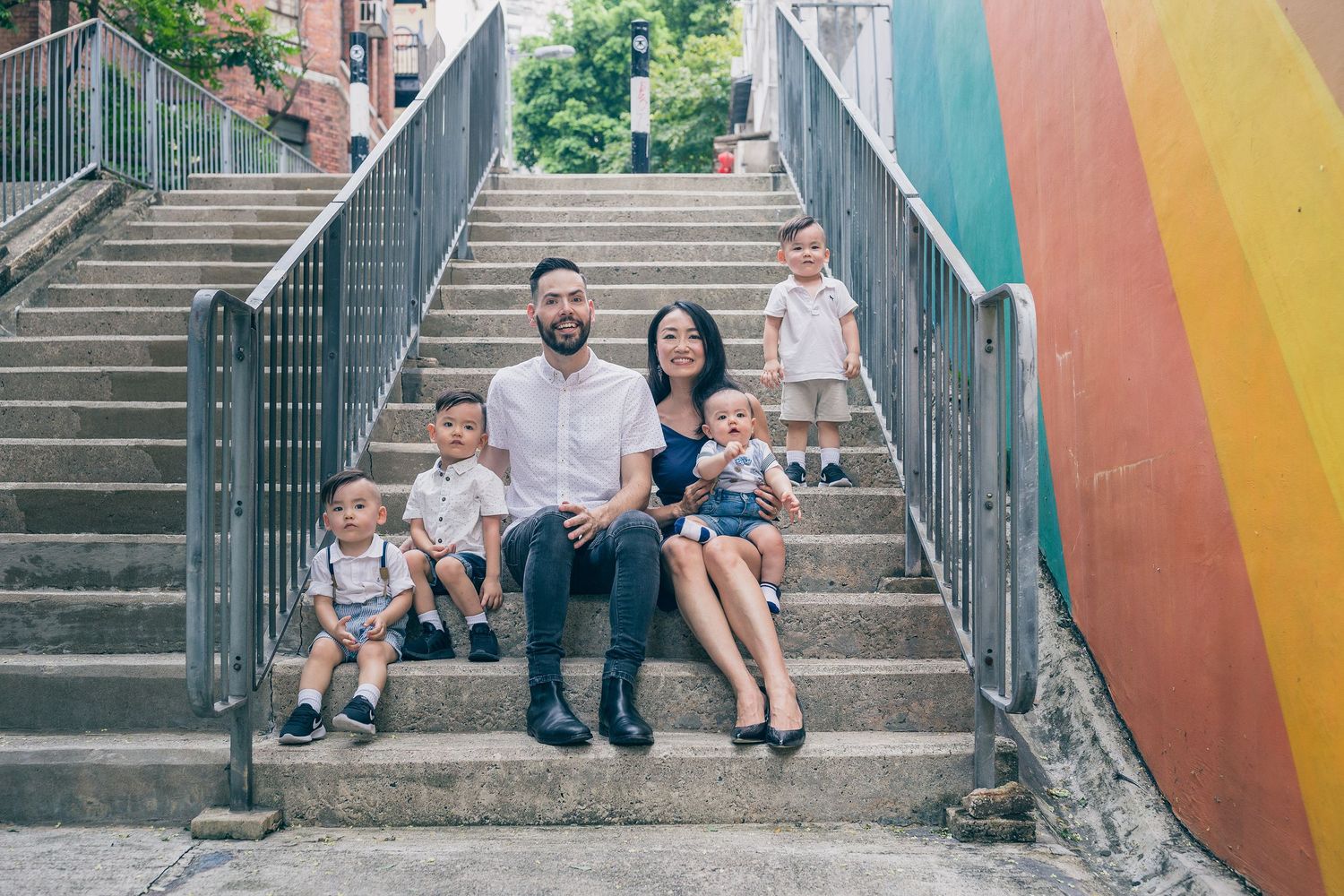 A cheerful family sits together on concrete steps next to a colorful wall on a sunny afternoon.