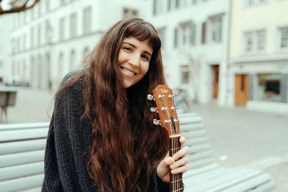 Personal branding an lifestyle photography: Young female musician sitting on a bench in Winterthur holding a guitar, smiling to the camera.