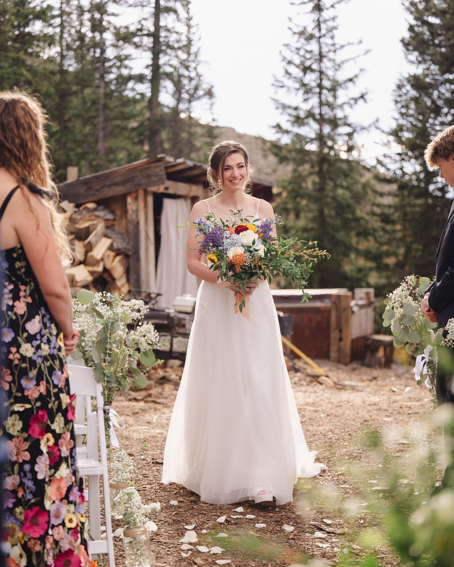 A bride in a flowing white dress holds a vibrant wildflower bouquet during an outdoor mountain wedding ceremony.