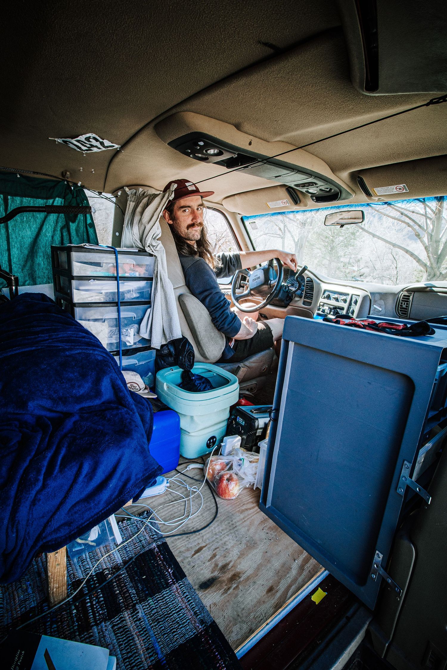 Interior view of van life with someone at the wheel surrounded by camping gear and electronics in a cluttered space.