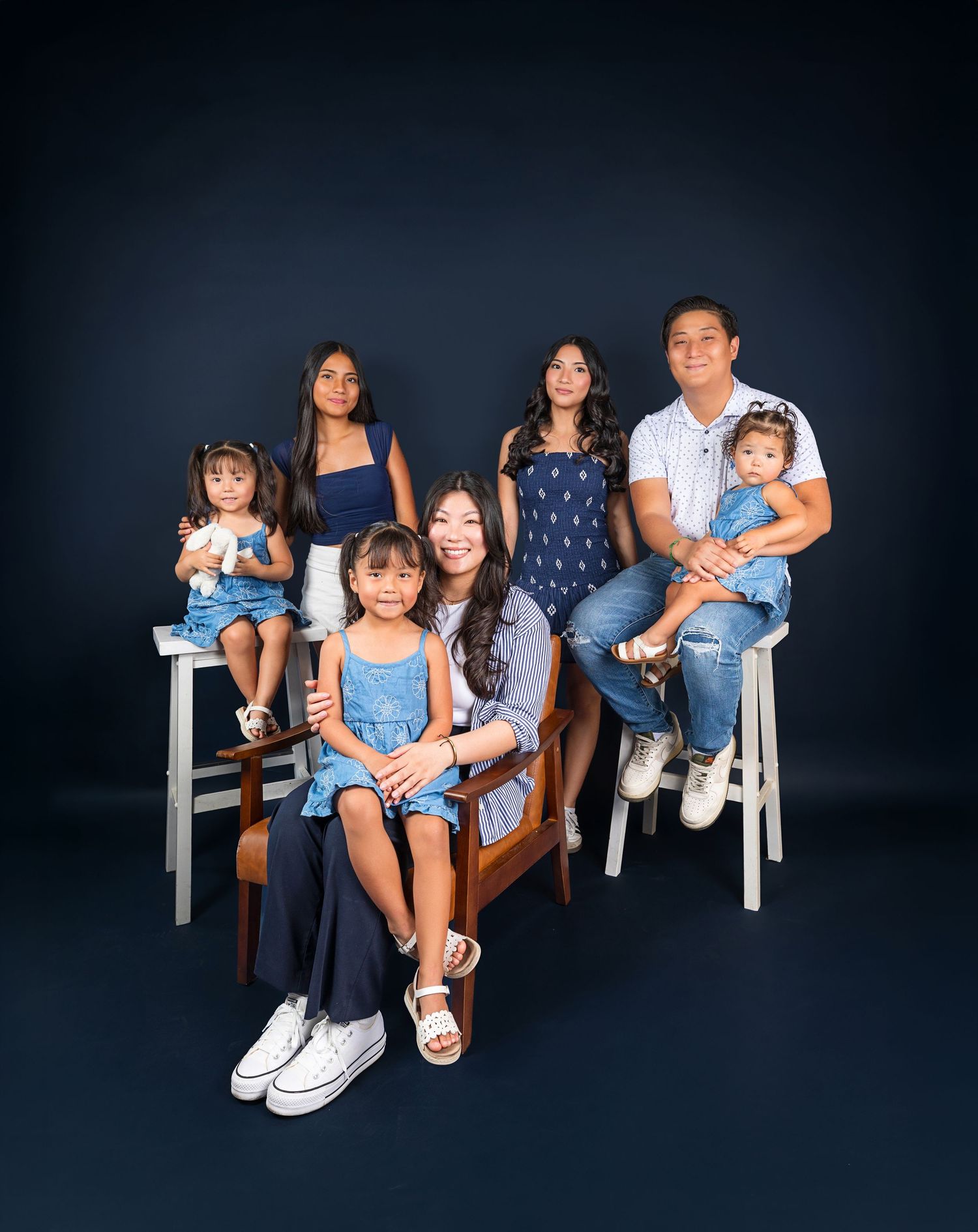Family portrait with blue color theme against dark background showing group seated on white stools in studio setting.