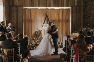 A wedding ceremony takes place inside a rustic barn venue with wooden walls and a draped fabric backdrop and floral decor.