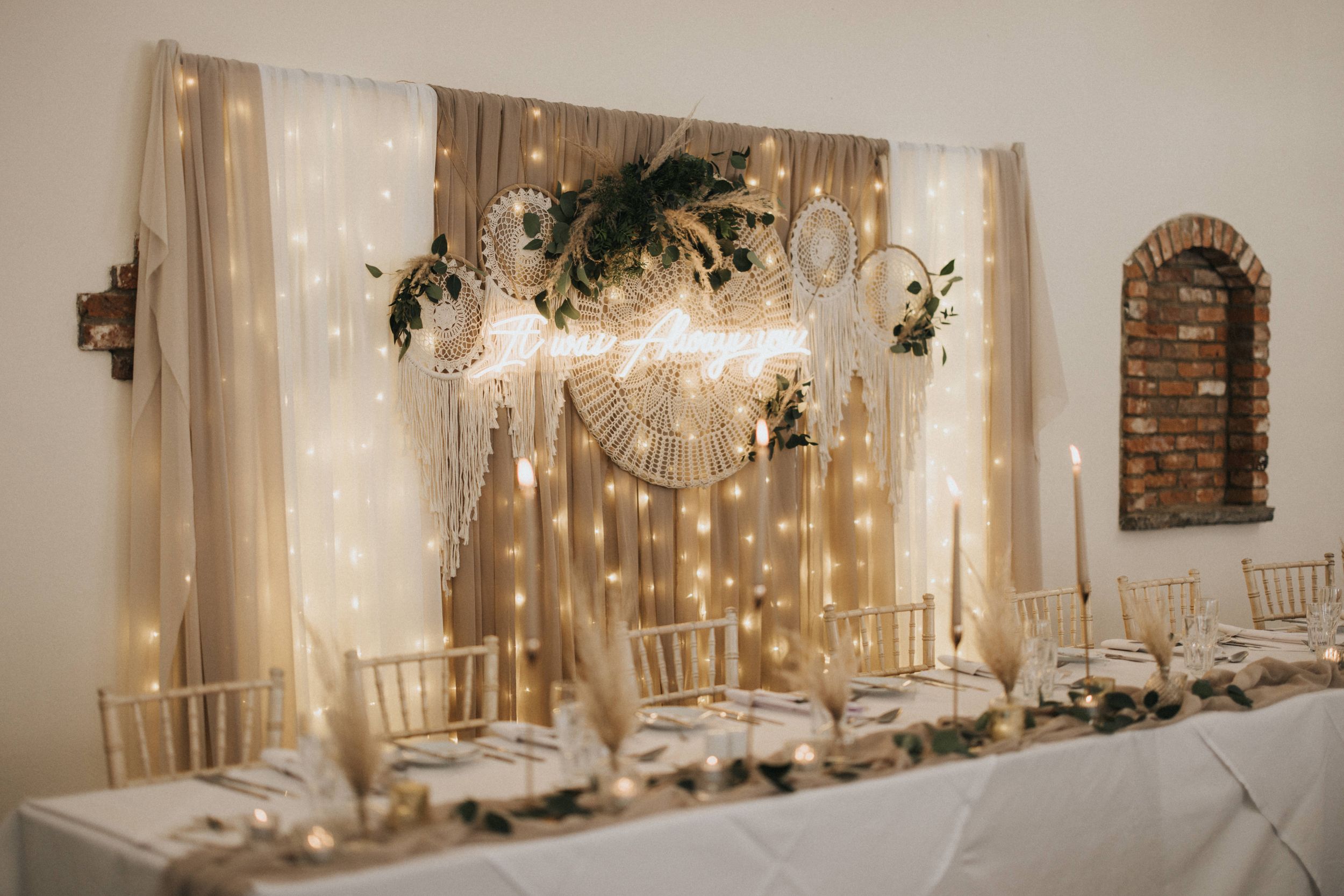Wedding venue decoration with warm twinkle lights, sheer white drapes, exposed brick wall and rustic wooden tables.