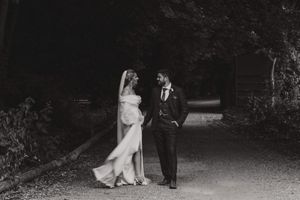 A couple in wedding attire stands together on a dark garden path in black and white photography.