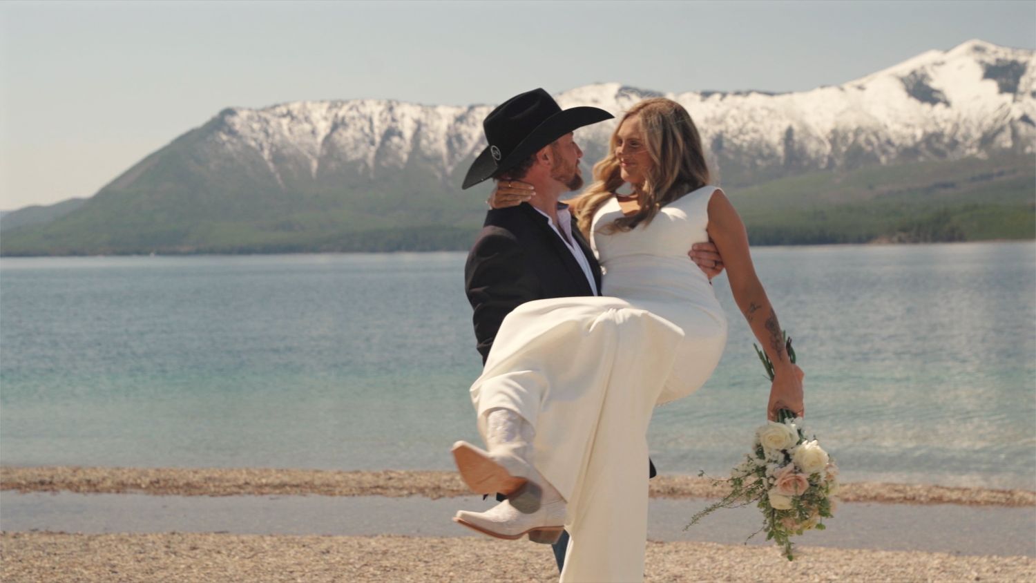 A romantic wedding couple shares a tender moment by a lake with snow-capped mountains in the background.