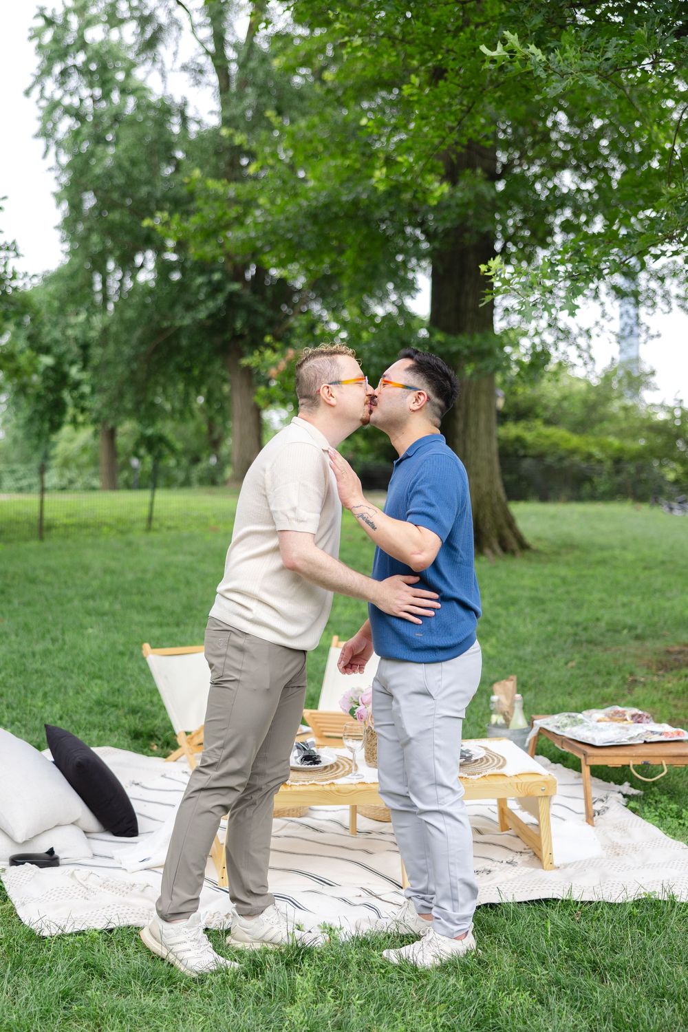 Two men kissing next to a romantic outdoor picnic setup with blankets and pillows in Central Park during a surprise proposal.