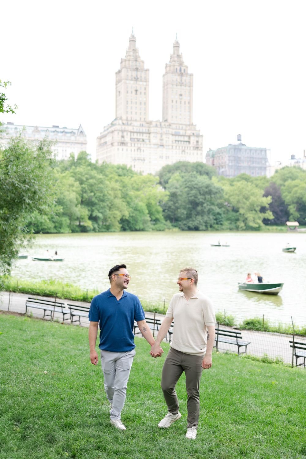 Two men walking together and laughing along the Central Park waterfront with Manhattan buildings in the background after their proposal