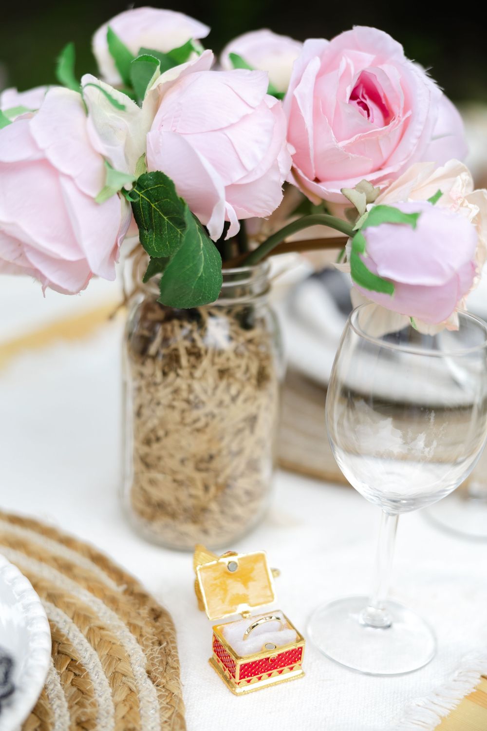Pink roses arranged in a mason jar with decorative details, wine glass and a ring box on a table setting.