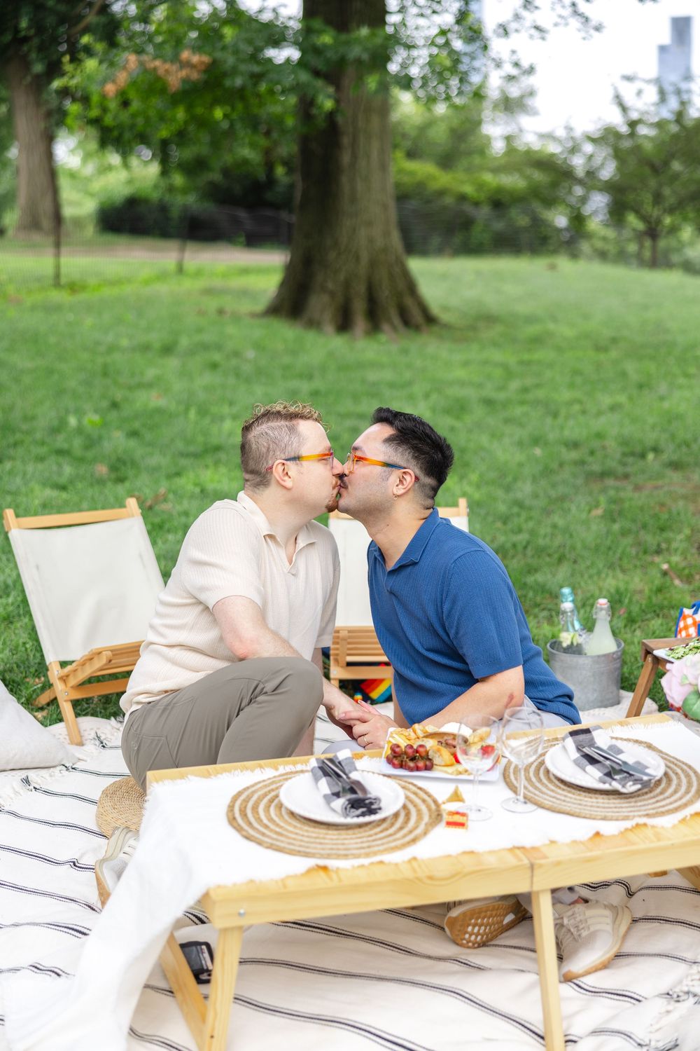 Two men kissing during an intimate picnic date in Central Park with romantic table setting and wine glasses.