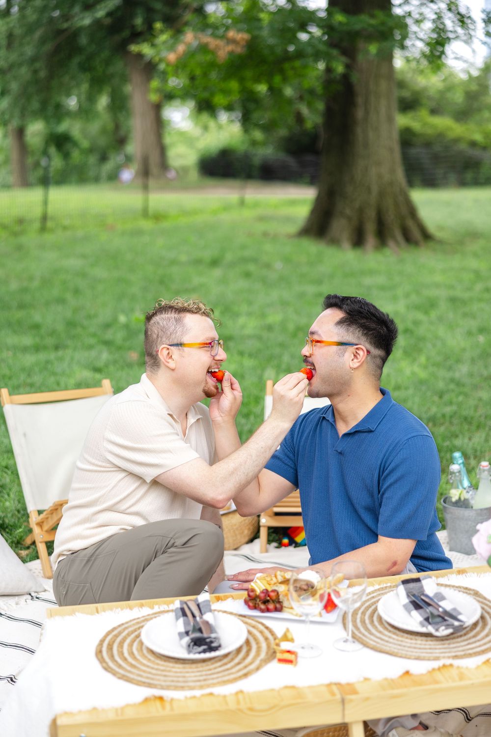 A newly engaged couple shares strawberries during a romantic outdoor picnic with table settings on the grass near trees.