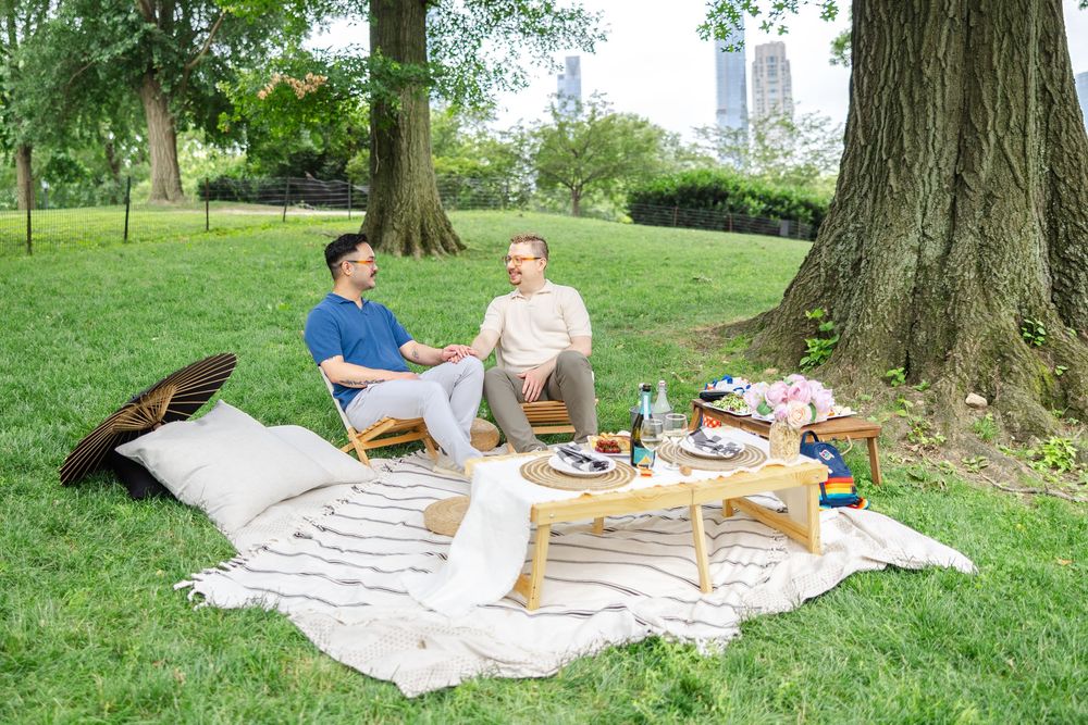 A cozy picnic scene with white blankets and wooden furniture arranged on grass under shady trees in Central Park during a surprise proposal.