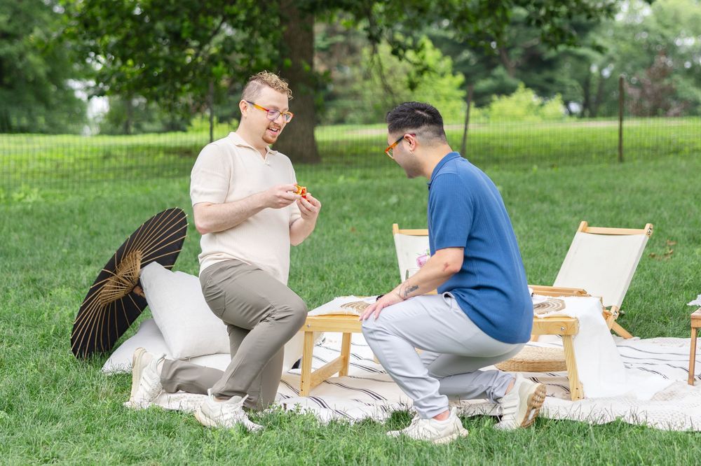 Two men kneeling on a white blanket during a proposal with a picnic in Cherry Hill of Central Park on a summer day. Photo by Joliette Marie.