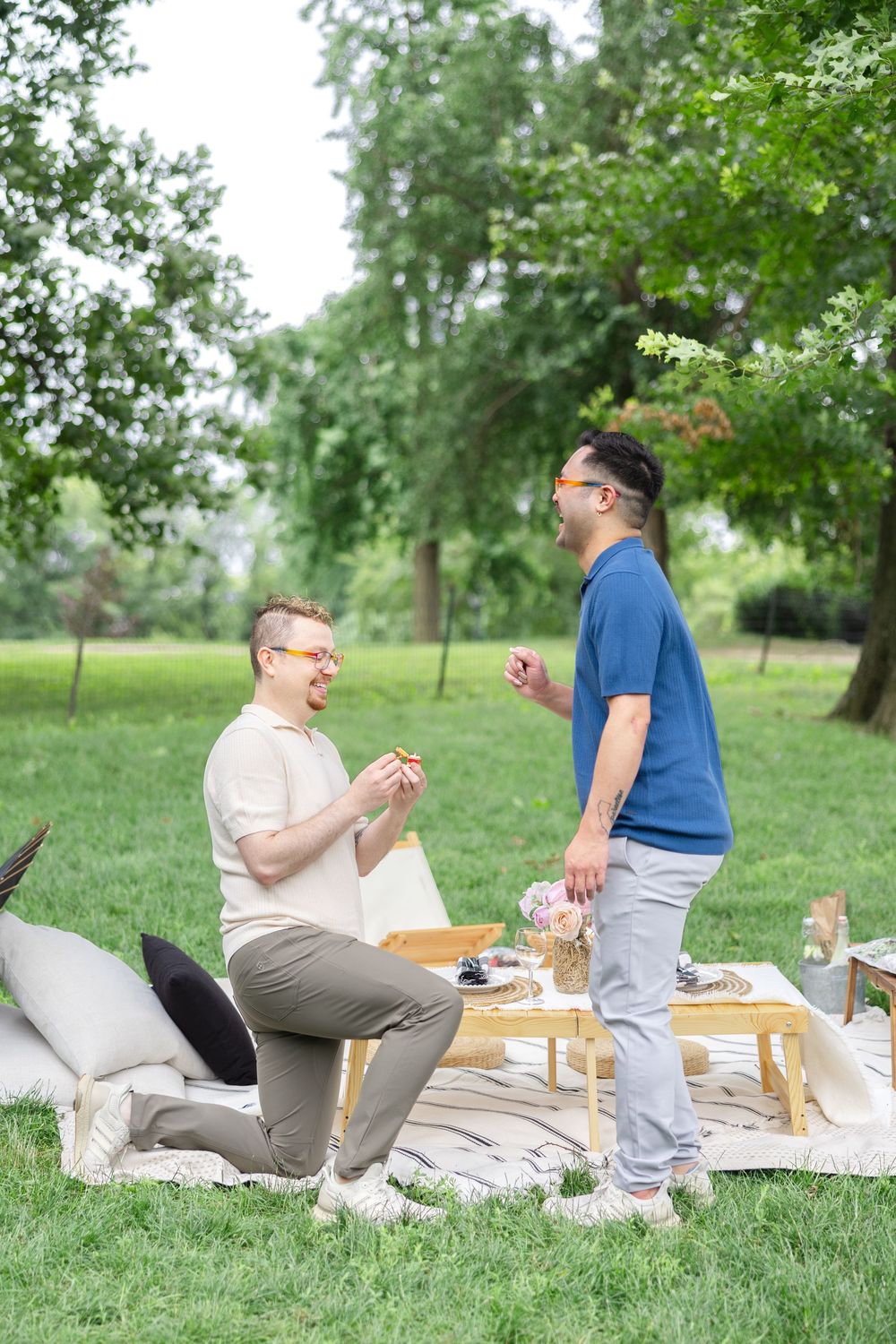 A romantic picnic proposal setup in a Central Park with glasses, blankets and a wooden table with a queer couple getting engaged.