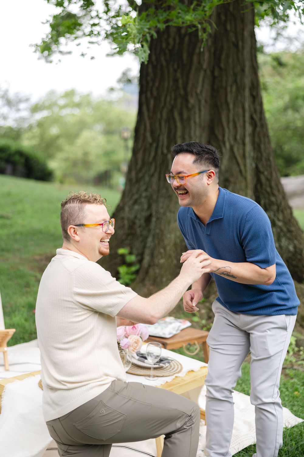 A gay couple laughs together during an outdoor picnic proposal in Central Park. Photo captured by Joliette Marie Photography.