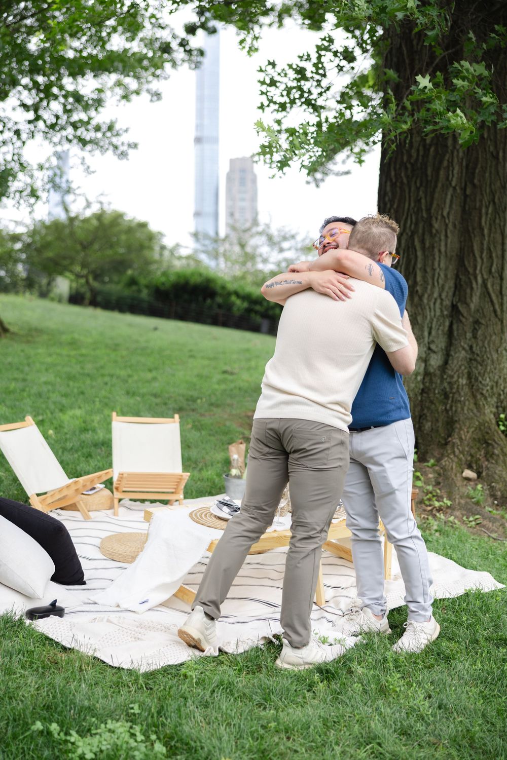 Two men hugging during a romantic picnic date on the grass near Central Park Lake with the iconic New York skyline visible.