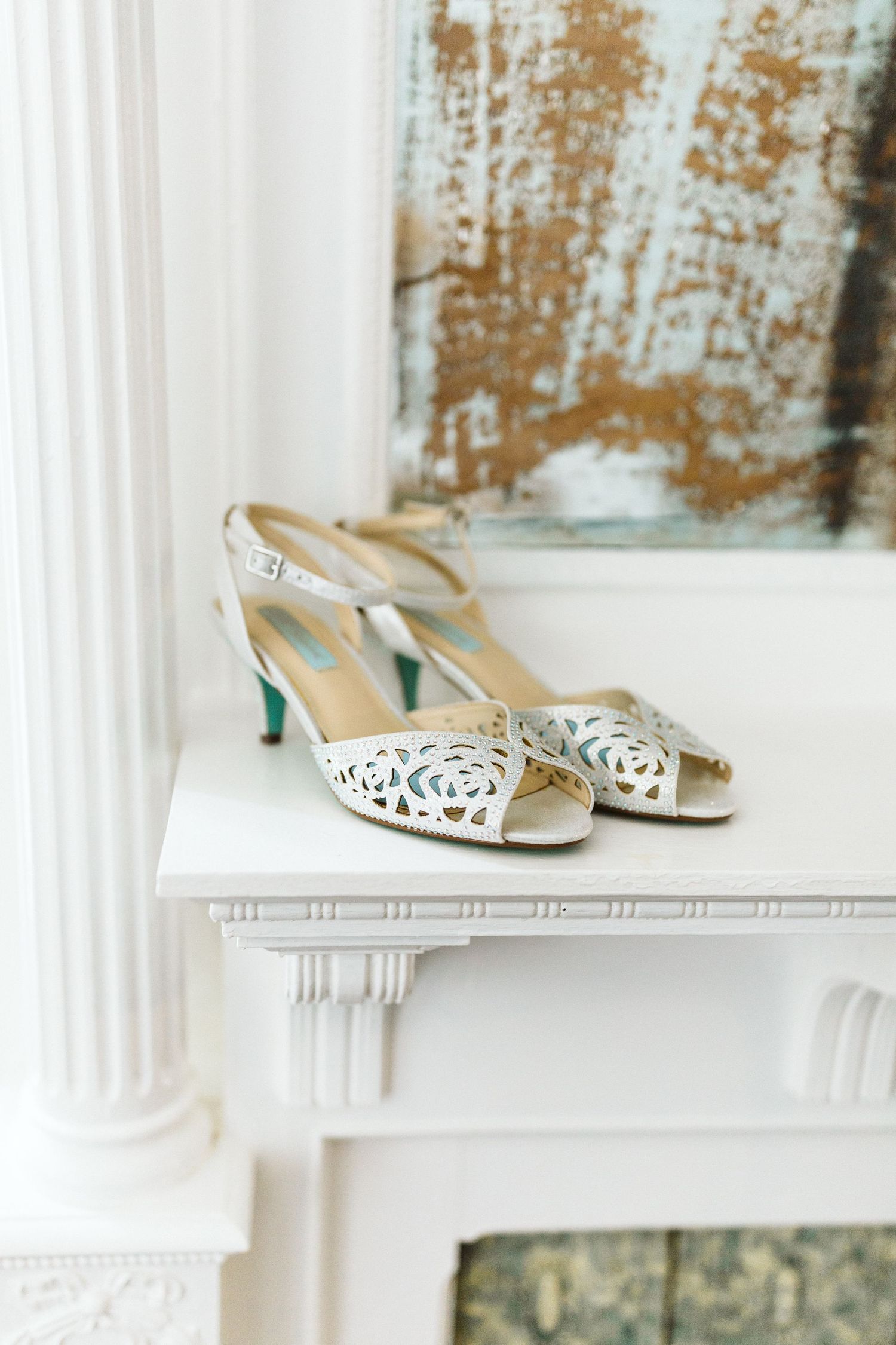 Elegant white cutout bridal shoes displayed on white shelf against rustic background.