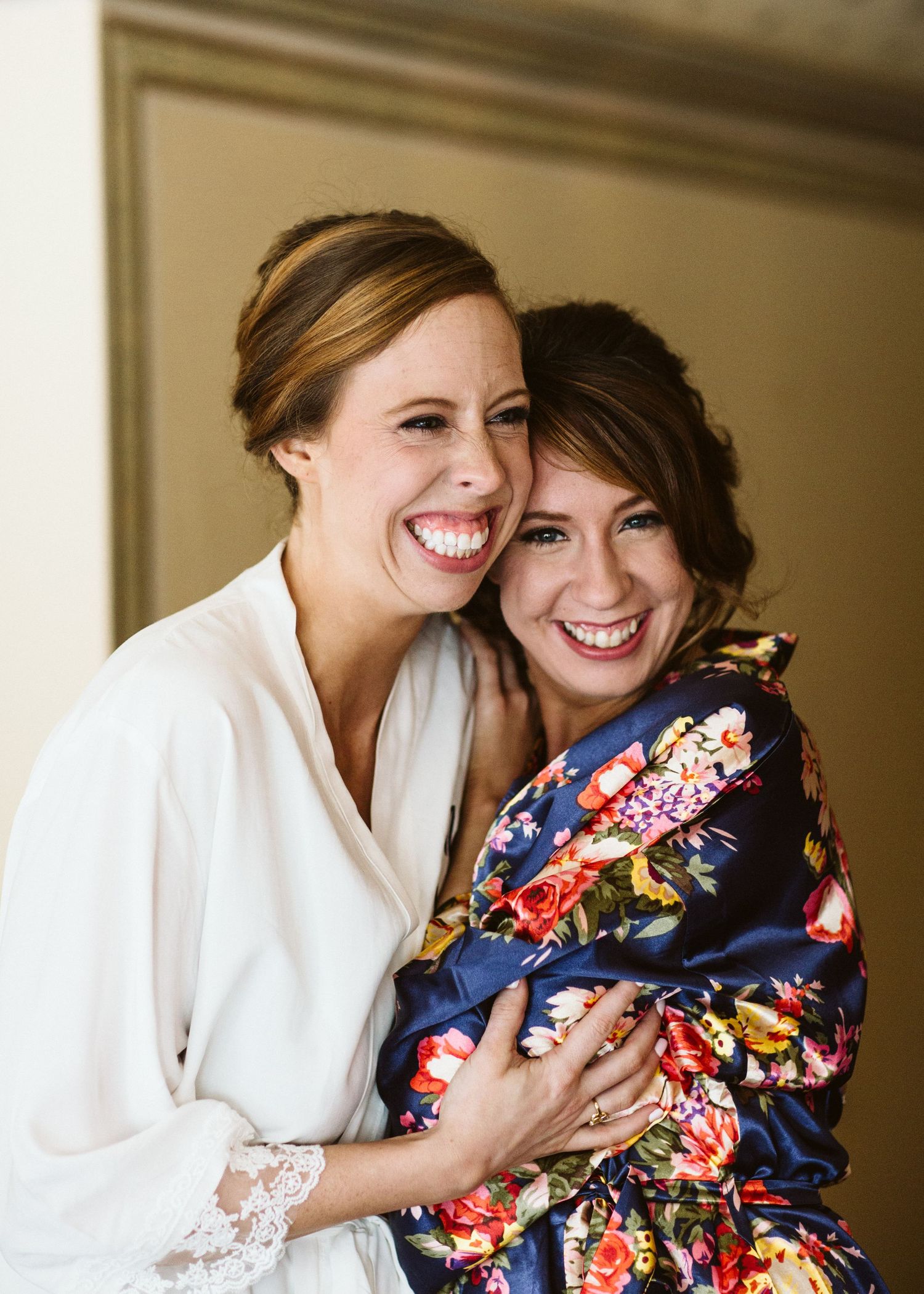 Two women share a joyful embrace while wearing a white robe and floral print robe.