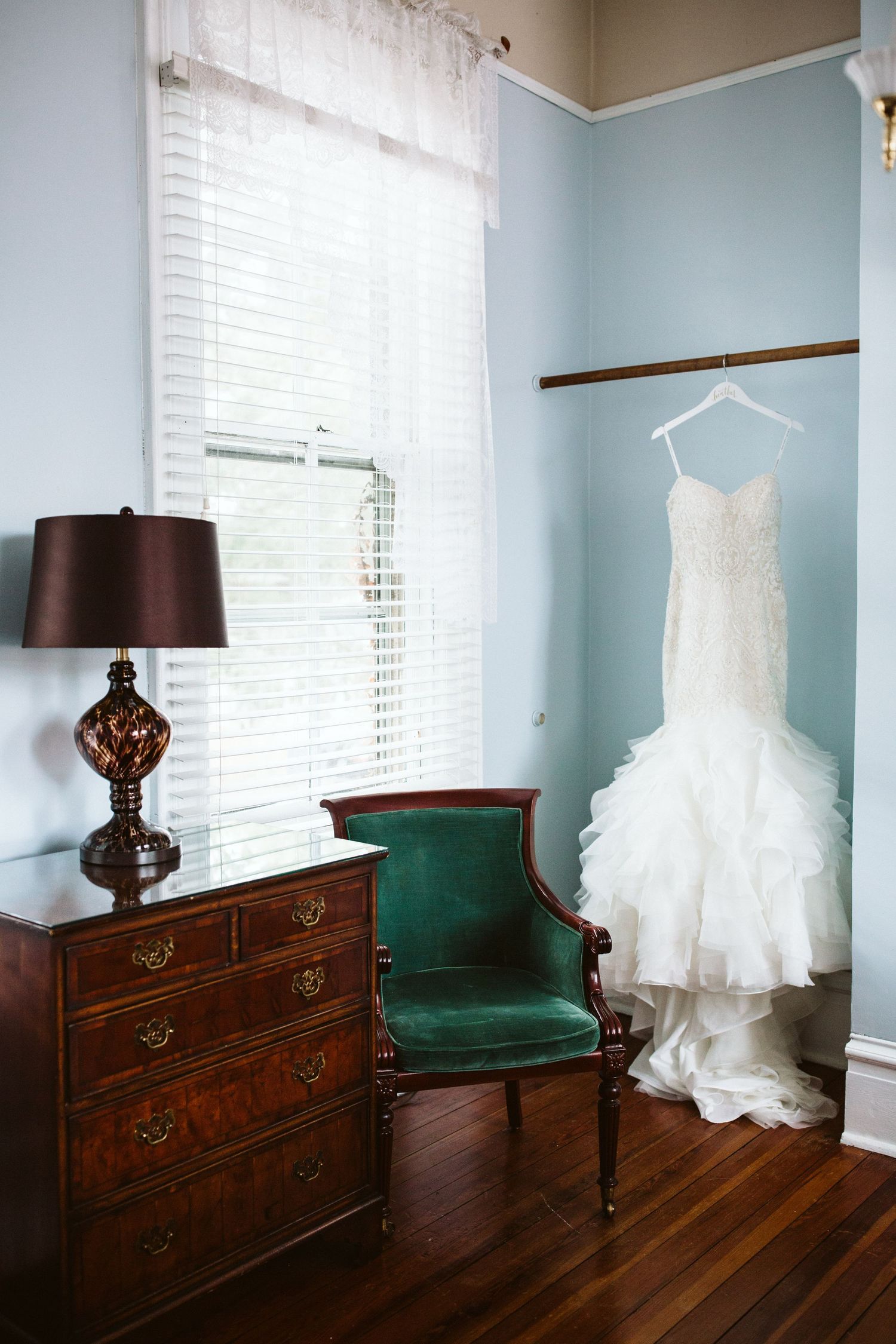White wedding dress displayed on hanger against light blue walls with dark wood dresser and emerald green chair.