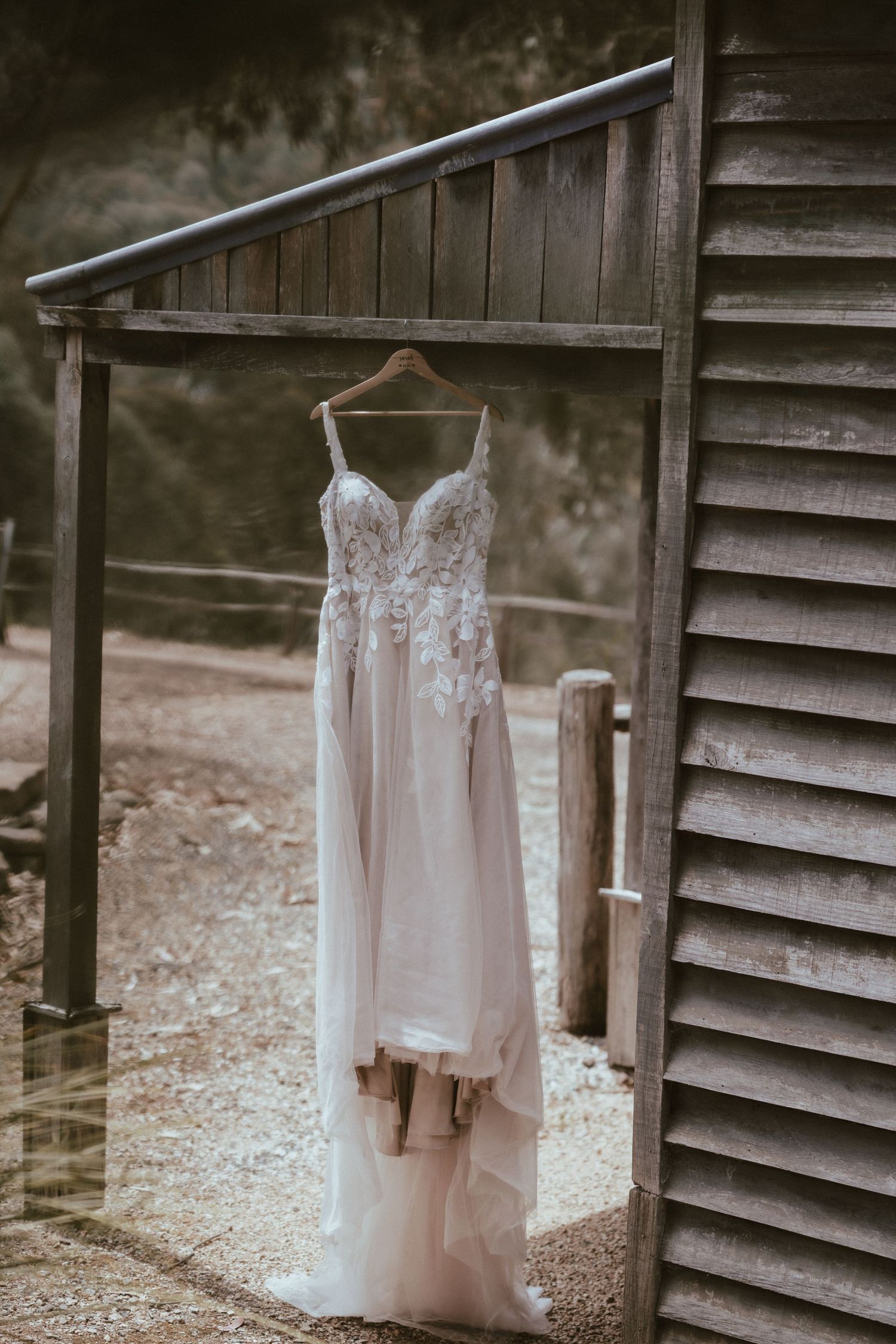 A delicate white lace wedding dress hangs on a wooden hanger against a rustic barn wall in soft lighting.