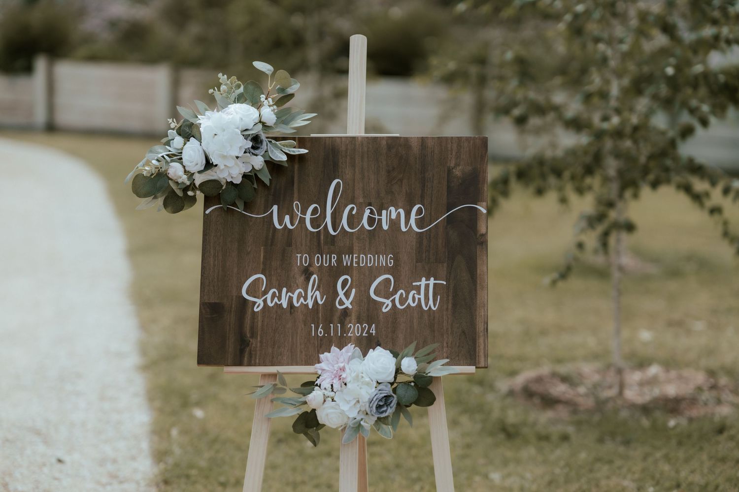 Rustic wooden wedding welcome sign decorated with white flowers and greenery against an outdoor background.