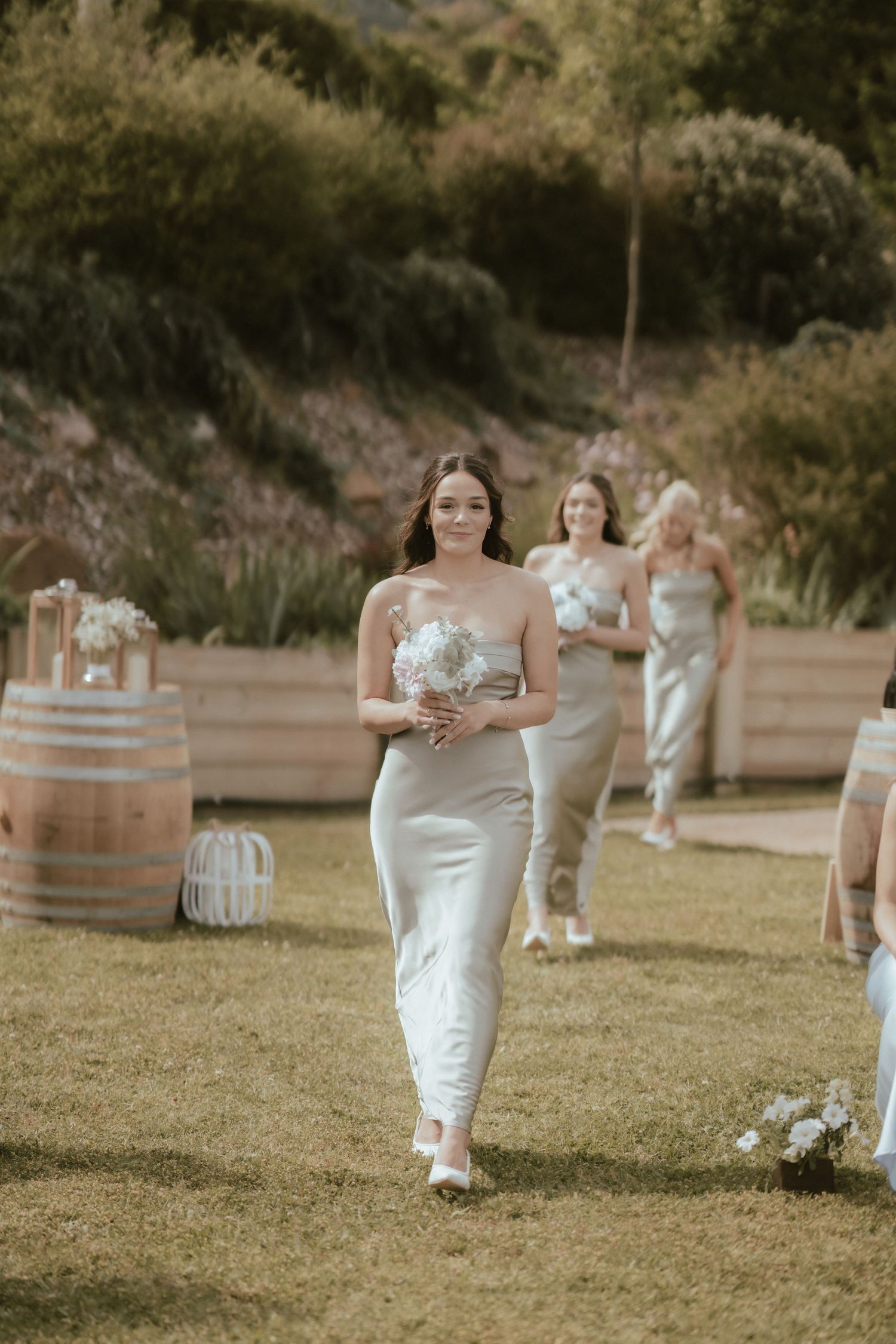 Bridesmaids in elegant white dresses walk down a grassy outdoor aisle with rustic wine barrels lining the ceremony space.