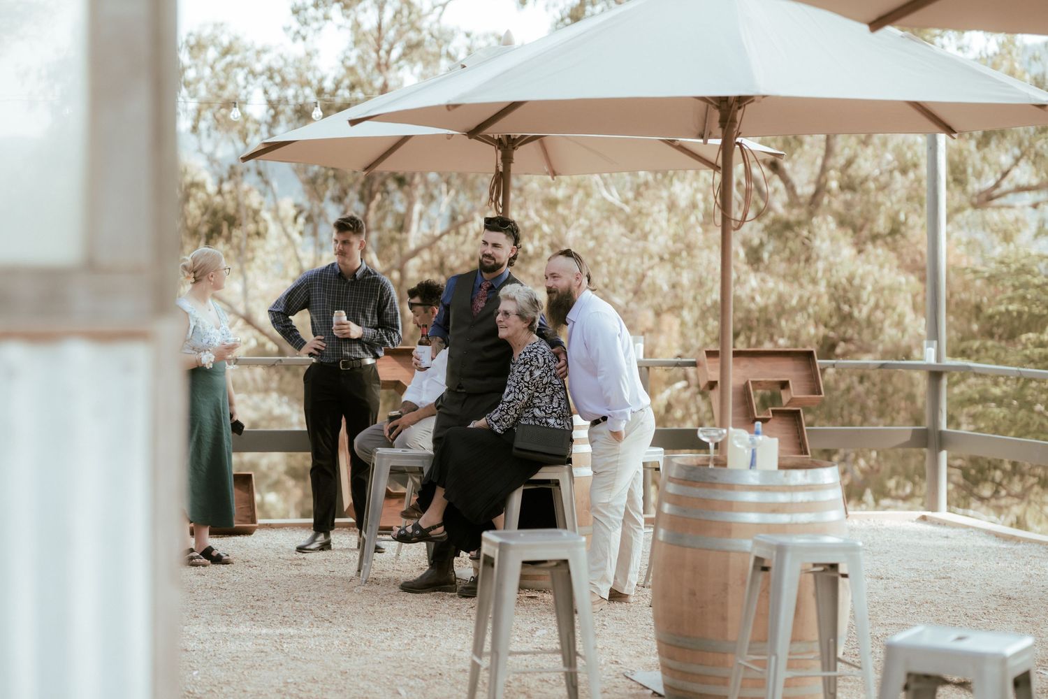 People gather at an outdoor winery tasting area with umbrellas and wine barrels on a sunny day.