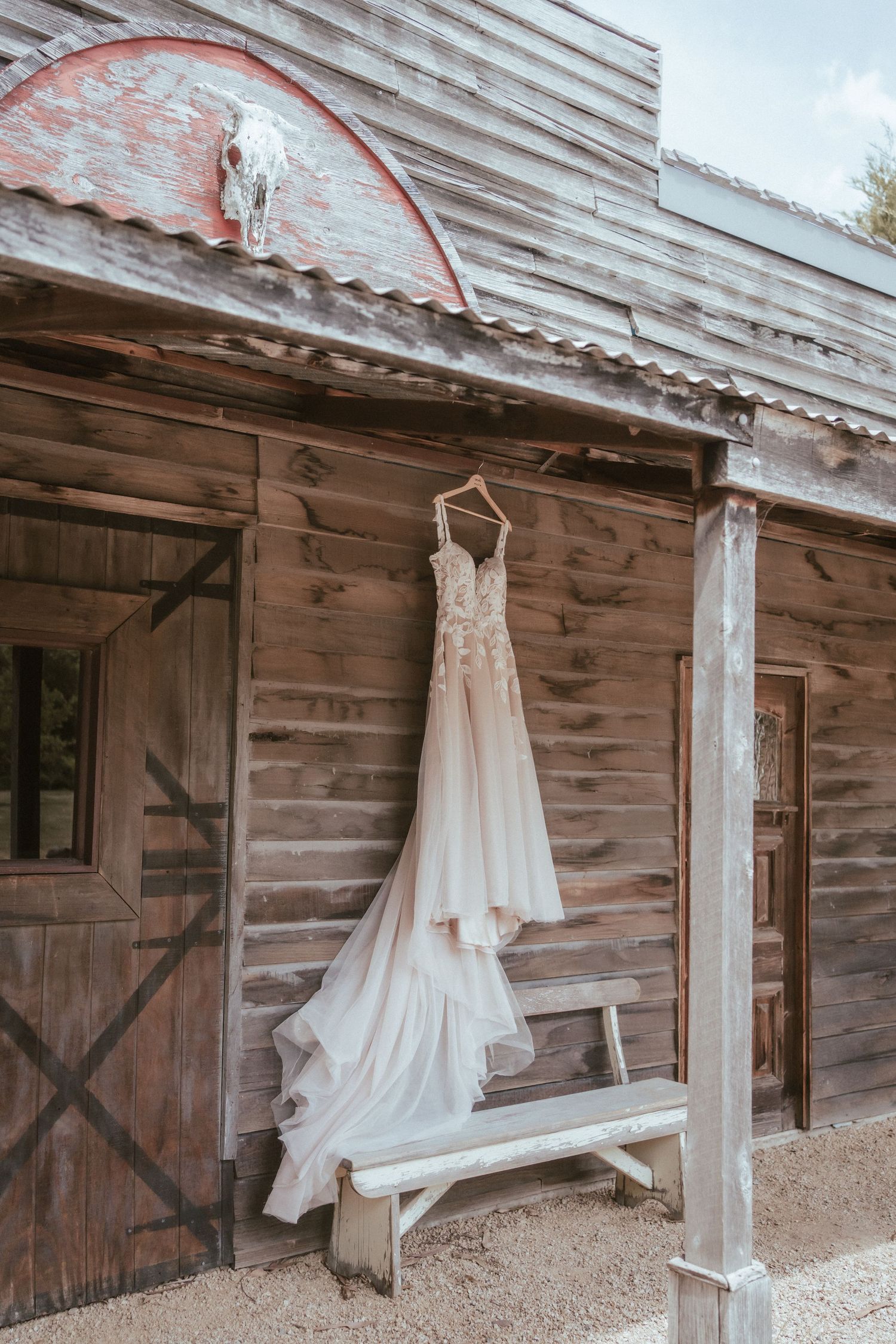 A white wedding dress hangs elegantly outside a rustic wooden barn with weathered siding and vintage doors.