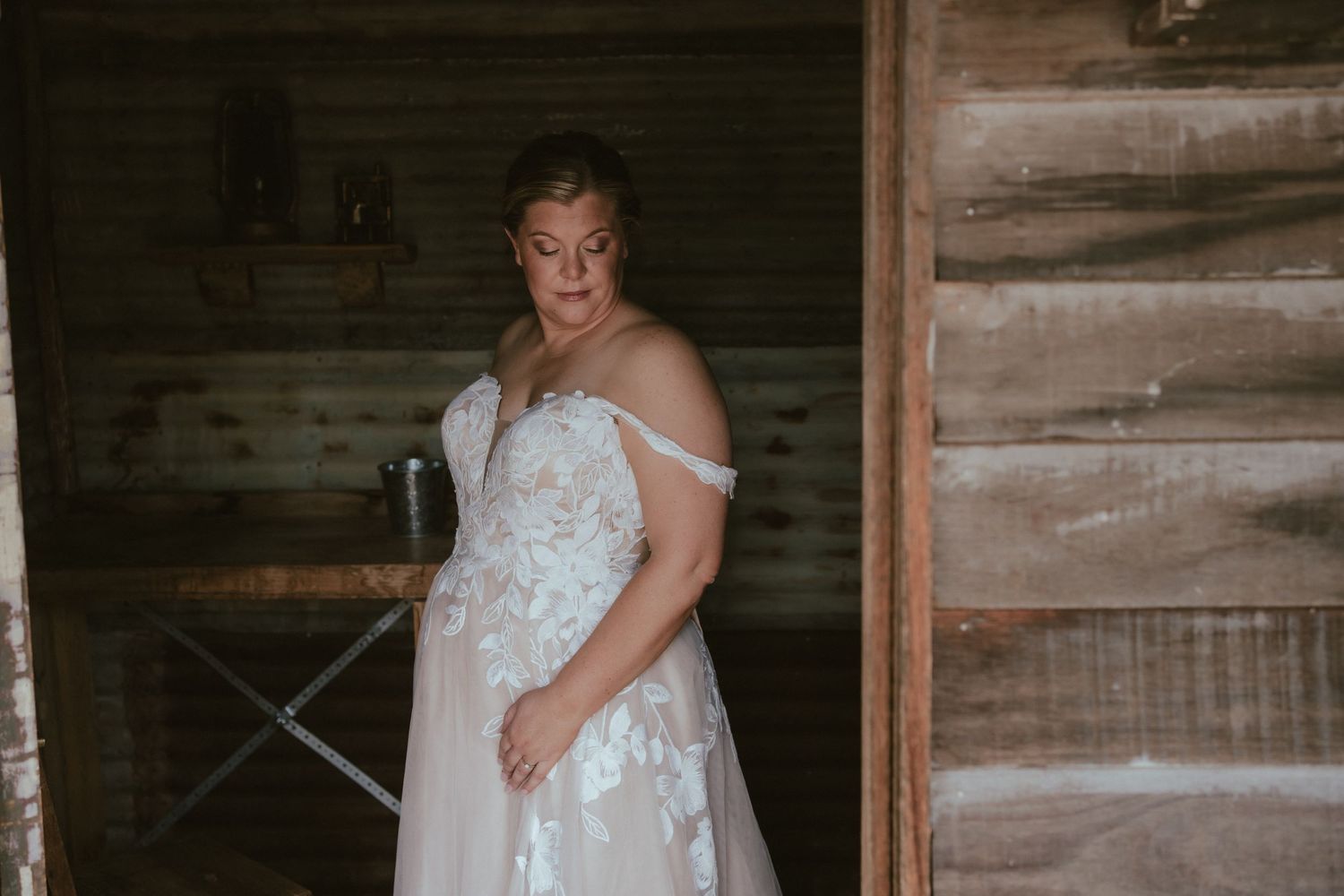 A person in an elegant white off-shoulder wedding dress with floral lace details poses inside a rustic wooden barn.