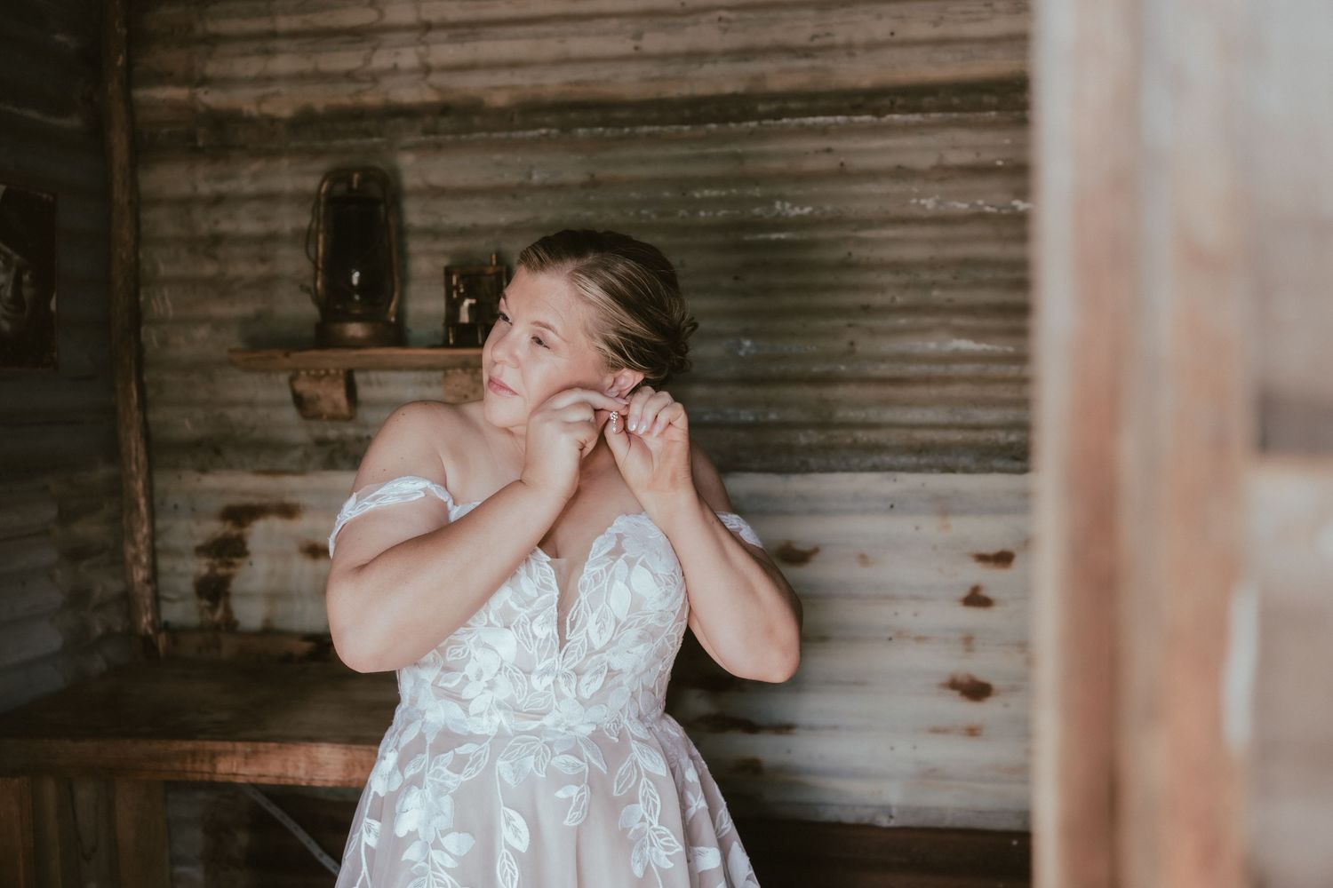 In a rustic barn setting, someone poses in an elegant white lace dress with off-shoulder detail.