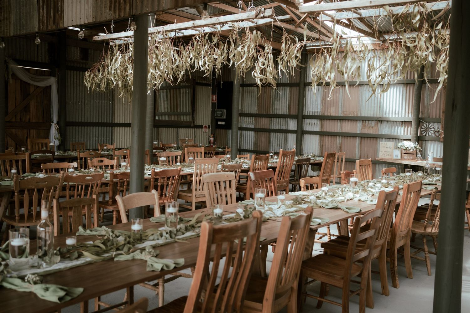 Long wooden dining tables set for a rustic wedding reception under dried floral installations in an industrial barn venue.