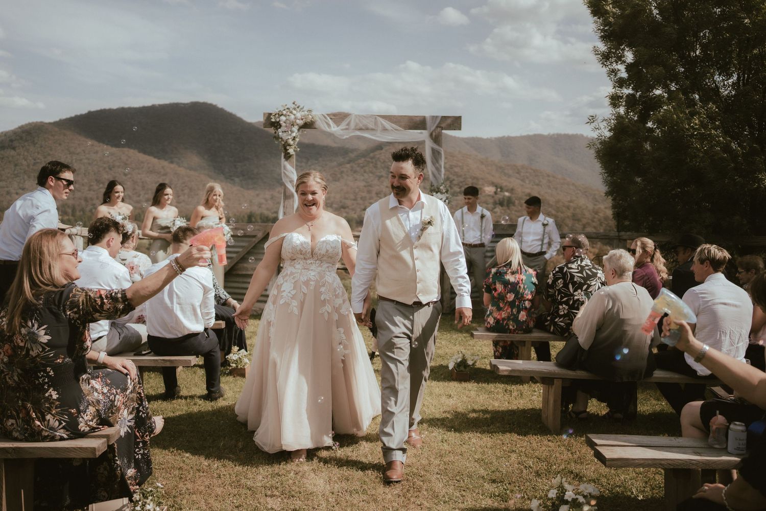 Outdoor mountain wedding ceremony with guests seated on rustic wooden benches during a sunny afternoon.