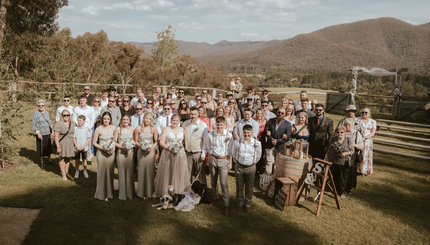 Large wedding party group photo captured in warm sunlight against a backdrop of mountains and trees at outdoor venue.
