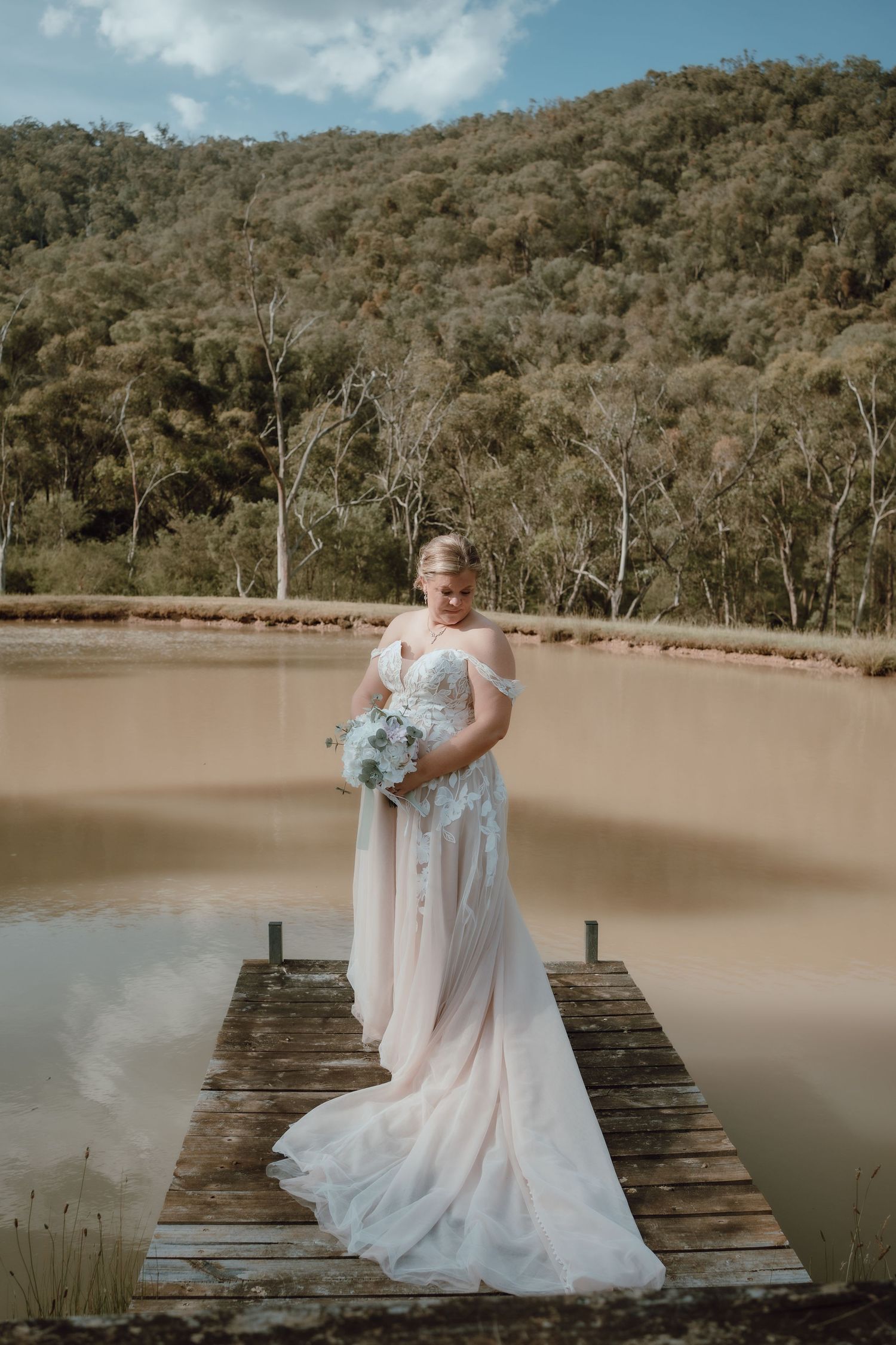 A white wedding gown with a flowing train photographed on a rustic wooden dock over calm water.