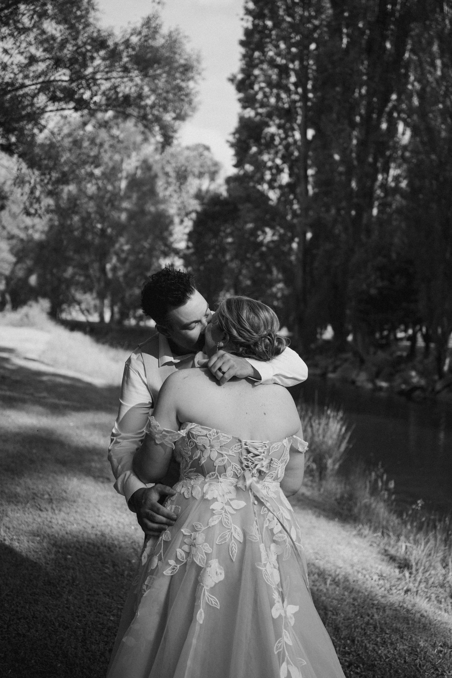 A tender moment captured in black and white by a lake with elegant wedding attire.