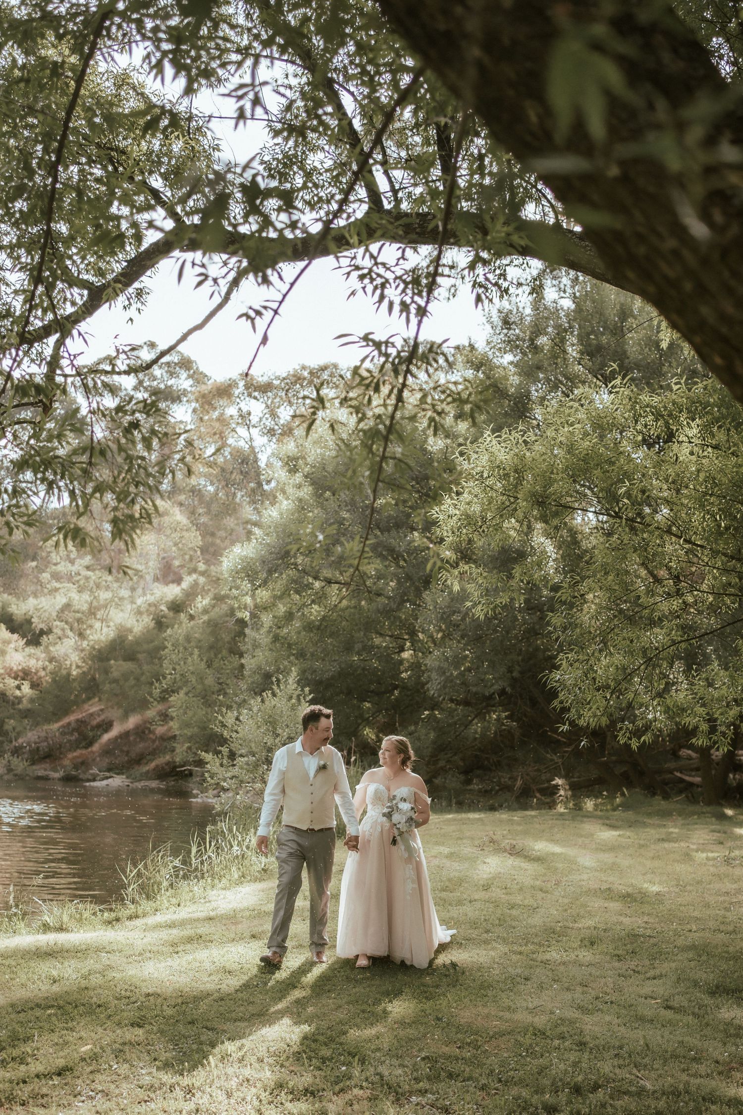 Couple shares intimate moment by a river under overhanging tree branches during their wedding day.