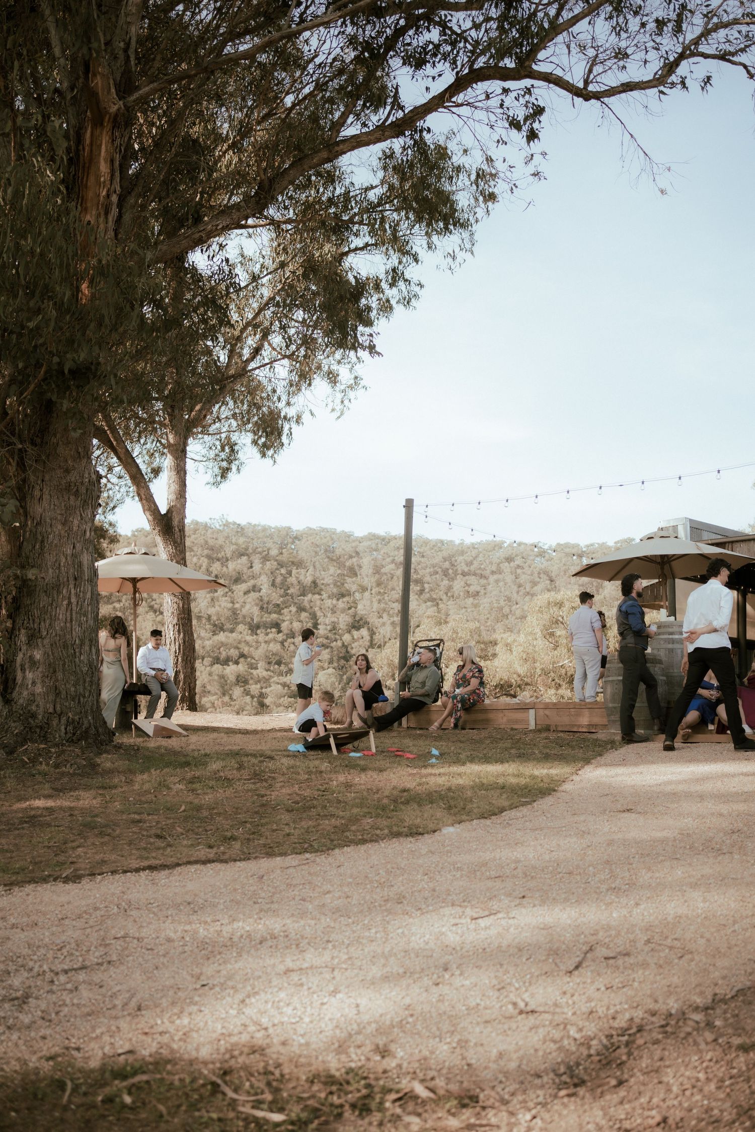 People gather at an outdoor venue with string lights and a large tree creating a rustic event atmosphere.