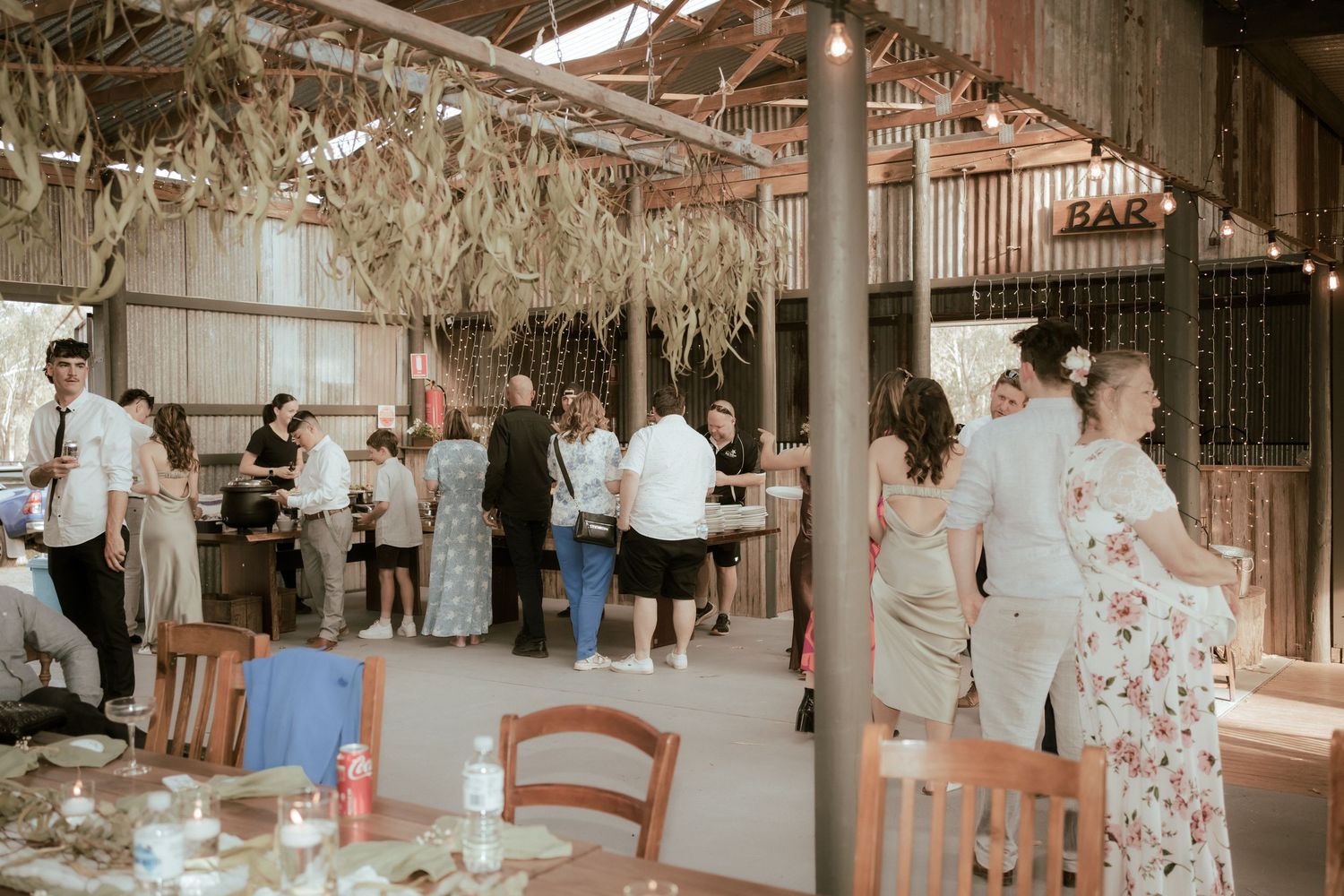 Guests mingle at a rustic wedding reception venue decorated with hanging dried flowers and string lights.