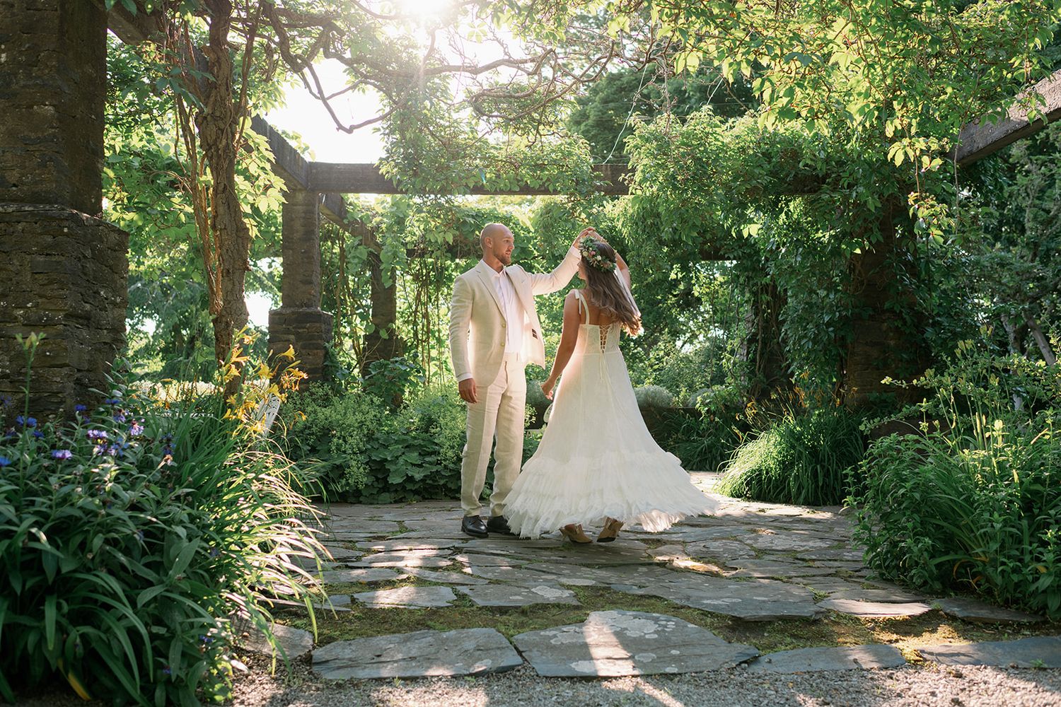 A couple walks along a sunlit garden path surrounded by lush greenery and blooming flowers on their wedding day.