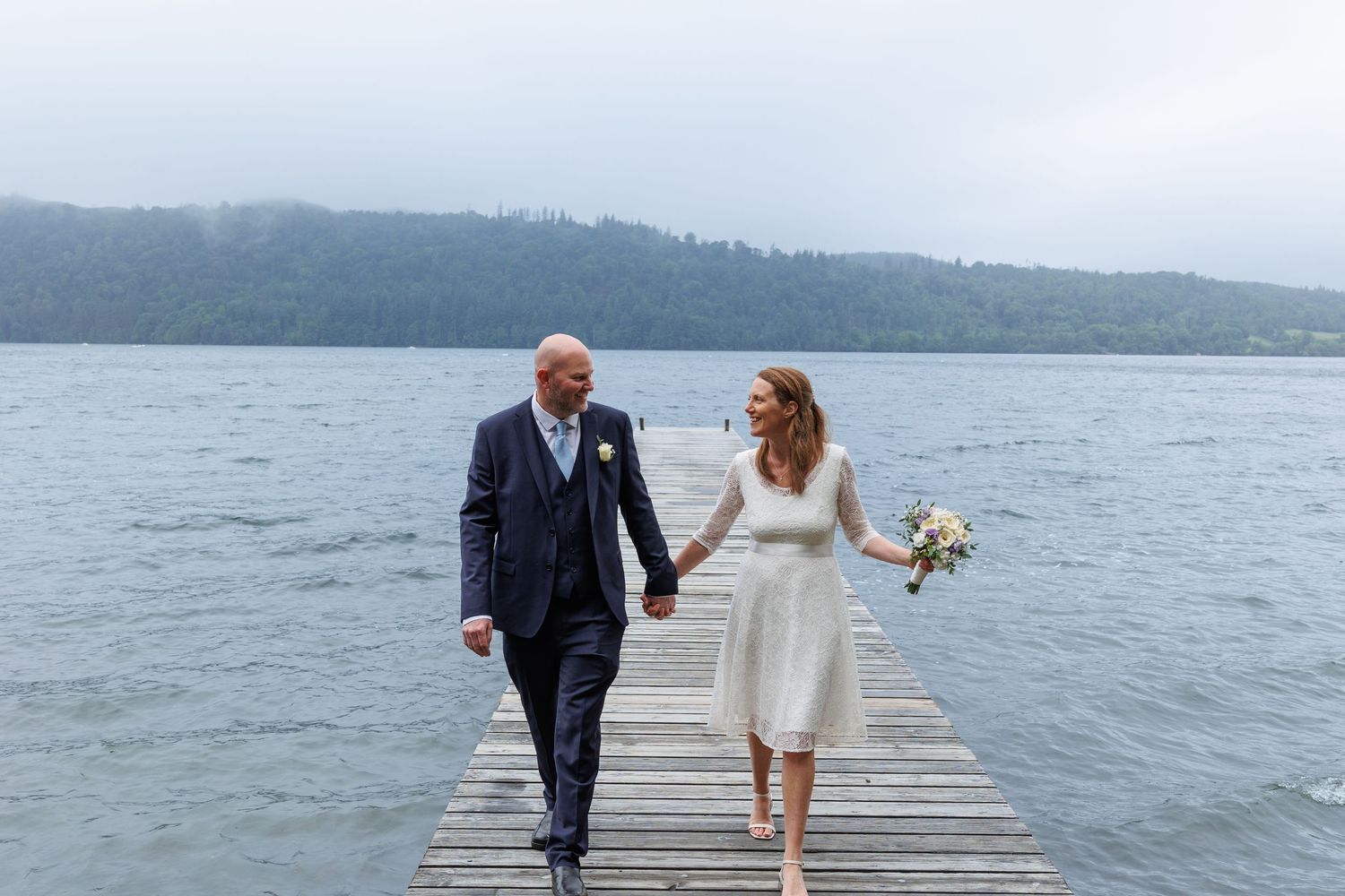 Couple walking on a wooden dock by a serene lake with mountains in the background.