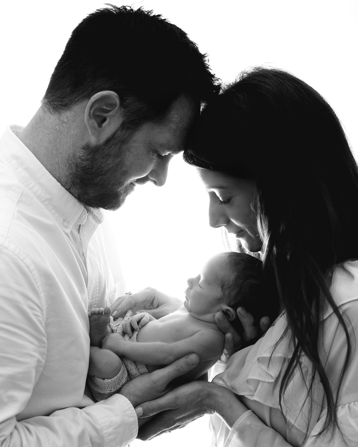 Black and white intimate family portrait, with parents in white clothes tenderly holding and gazing at their baby at a newborn session