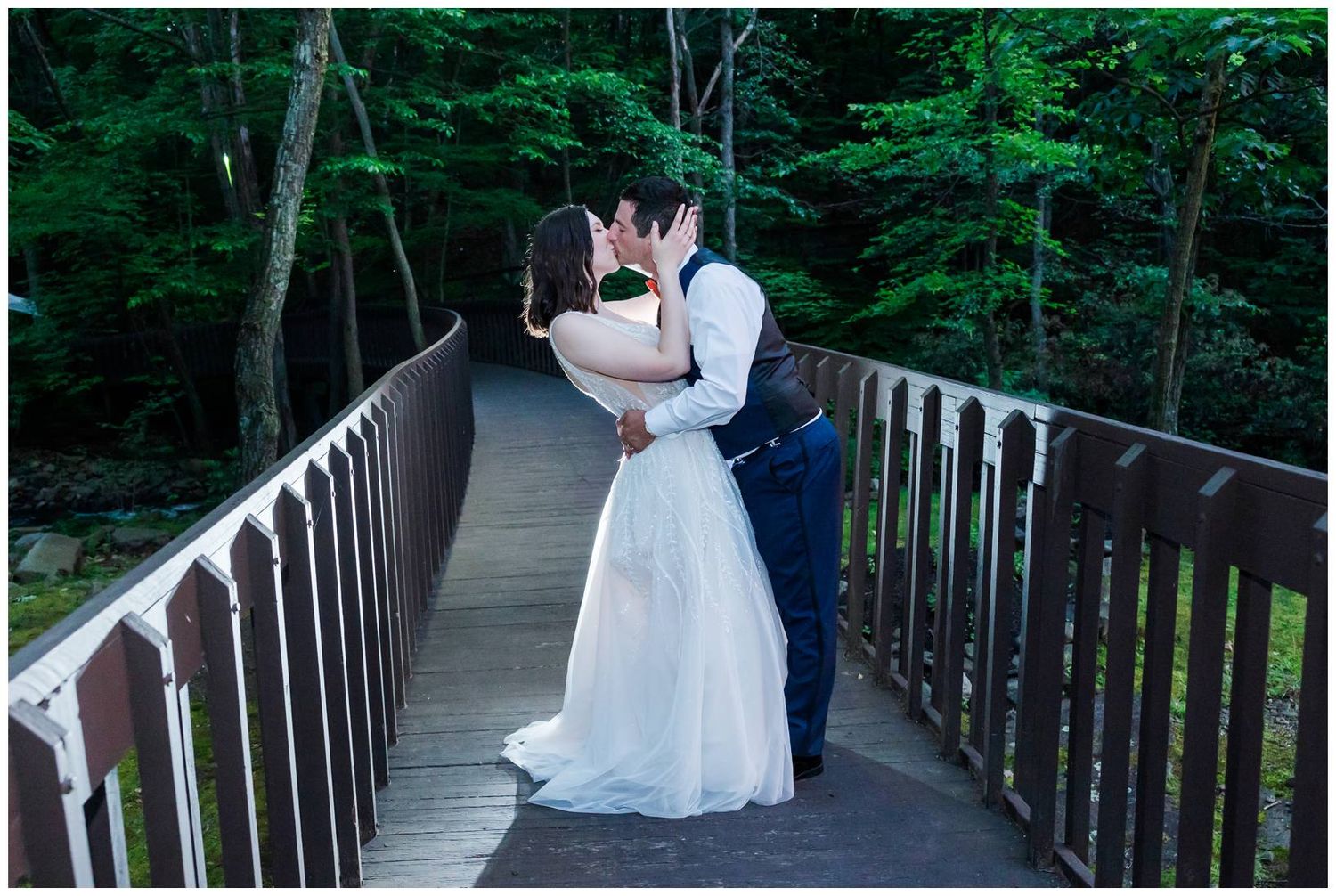 A couple in wedding attire shares an intimate moment on a wooden bridge surrounded by lush green forest.