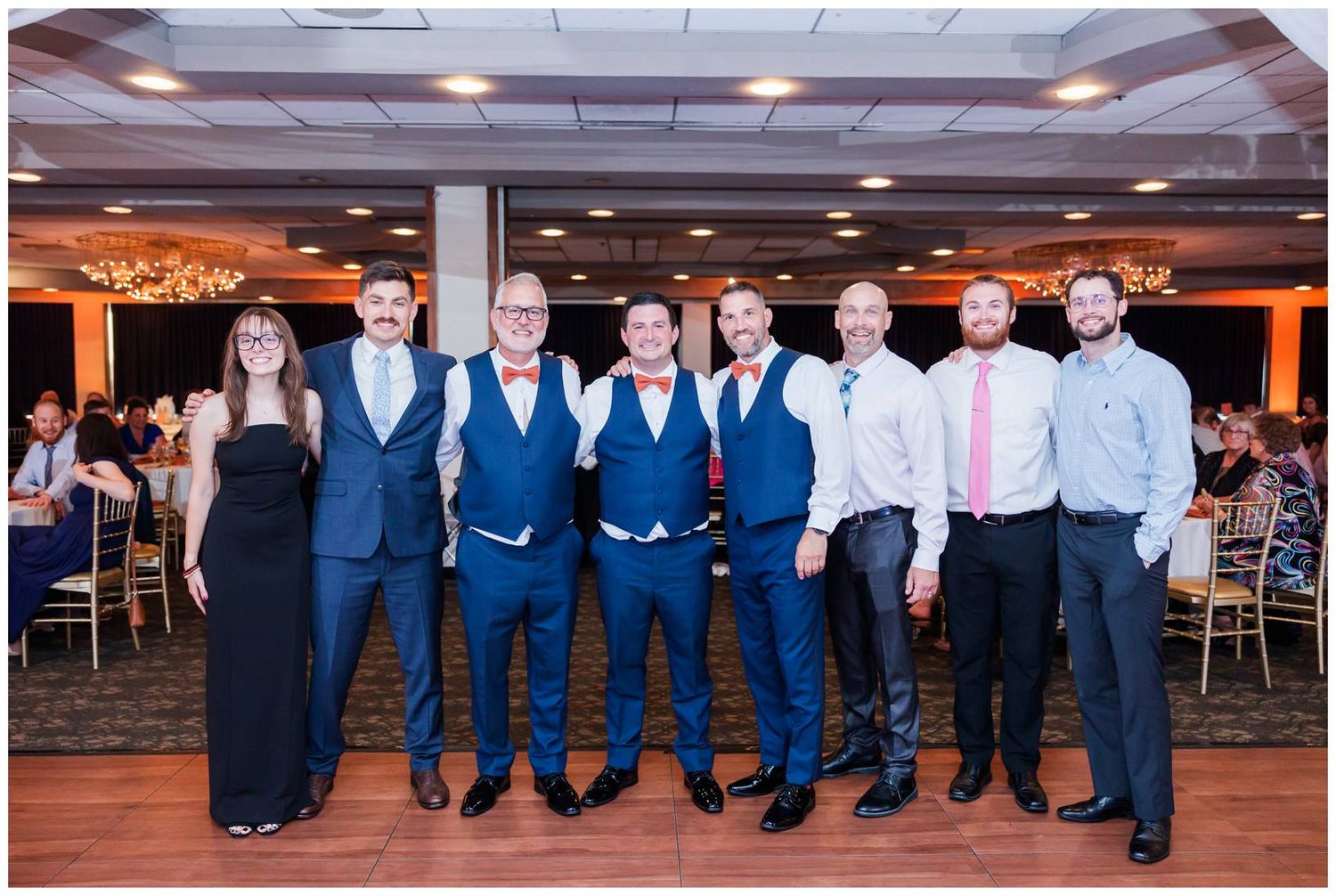A wedding party group poses together in a banquet hall with blue suits and formal attire against orange lighting.
