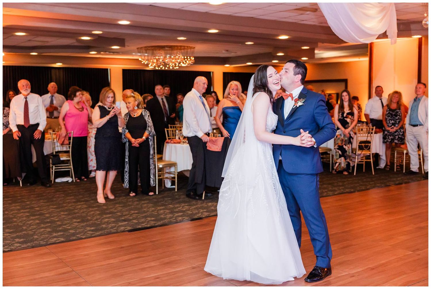 A couple shares their first dance as newlyweds on a wooden dance floor while guests watch from the reception venue.