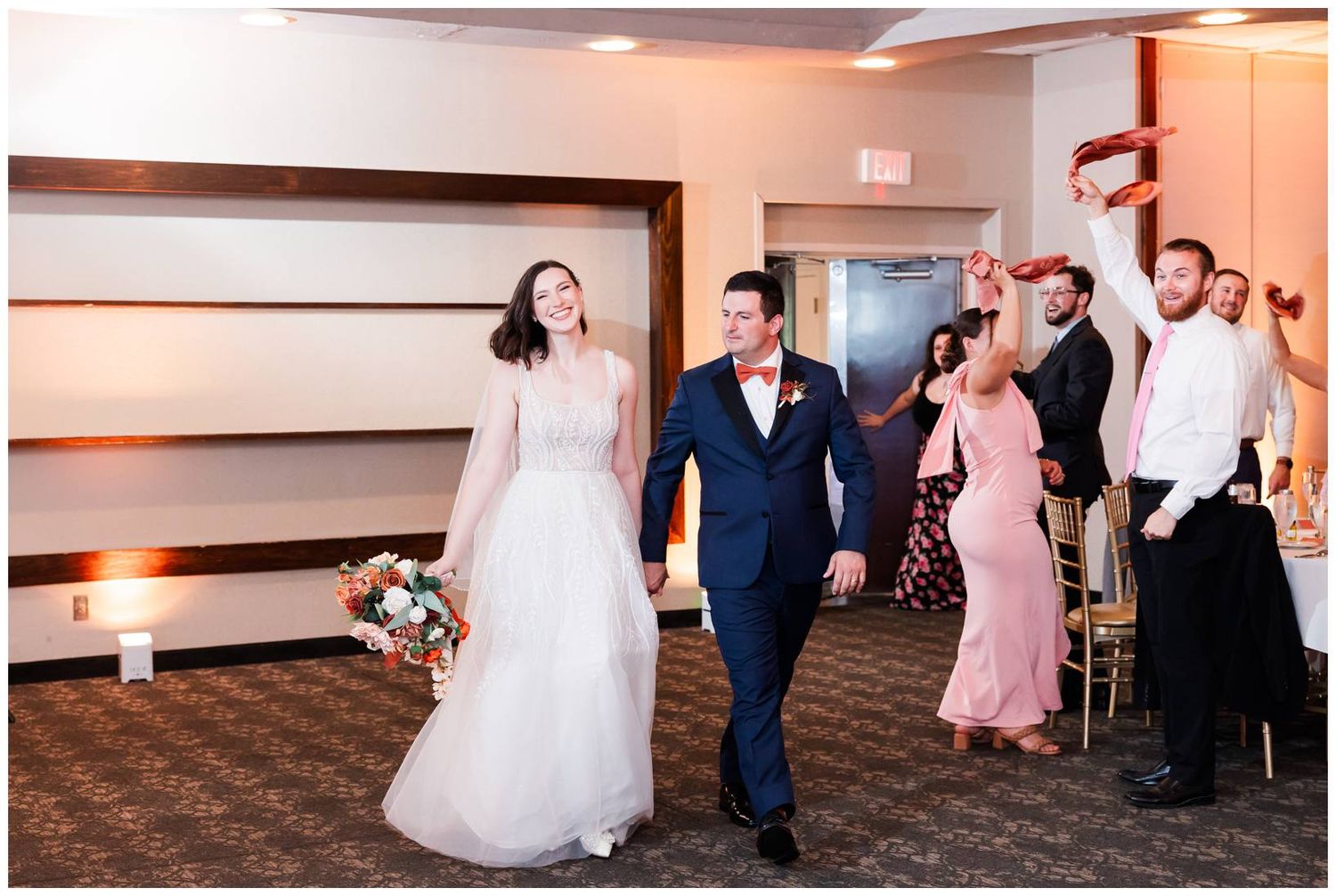 Wedding couple walking at reception with wedding party members cheering and celebrating behind them.