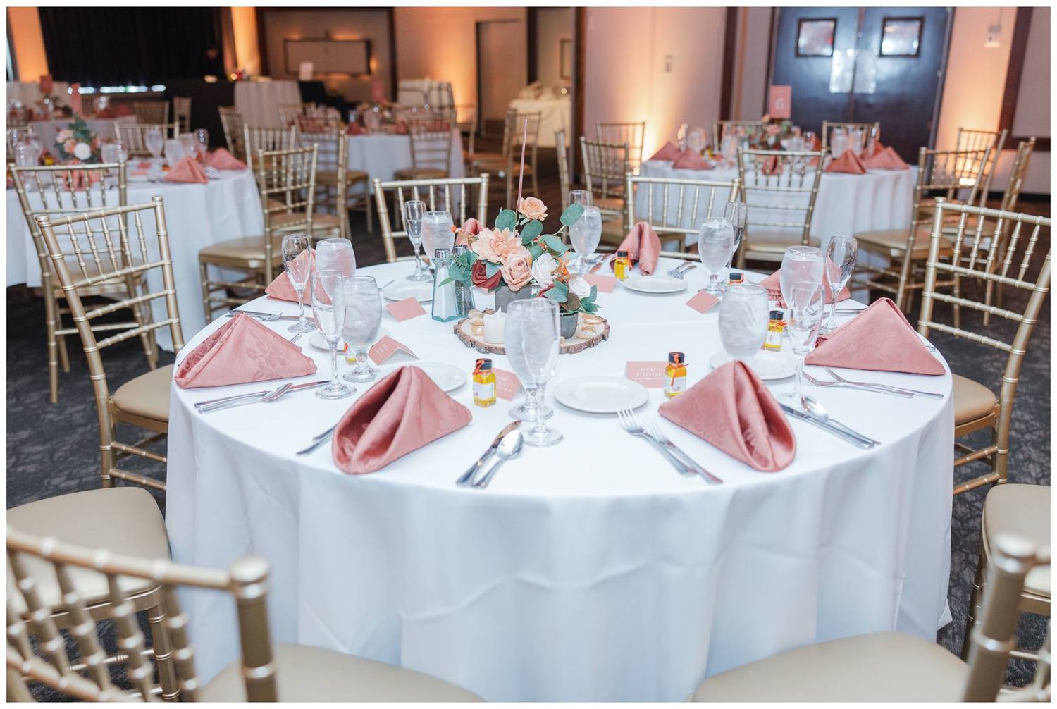 Elegant wedding reception table setting with white linens, rose gold napkins, and centerpieces in a ballroom venue.