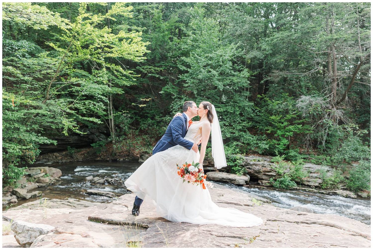 A couple in wedding attire embracing on a rocky outcrop surrounded by lush green forest and flowing water.